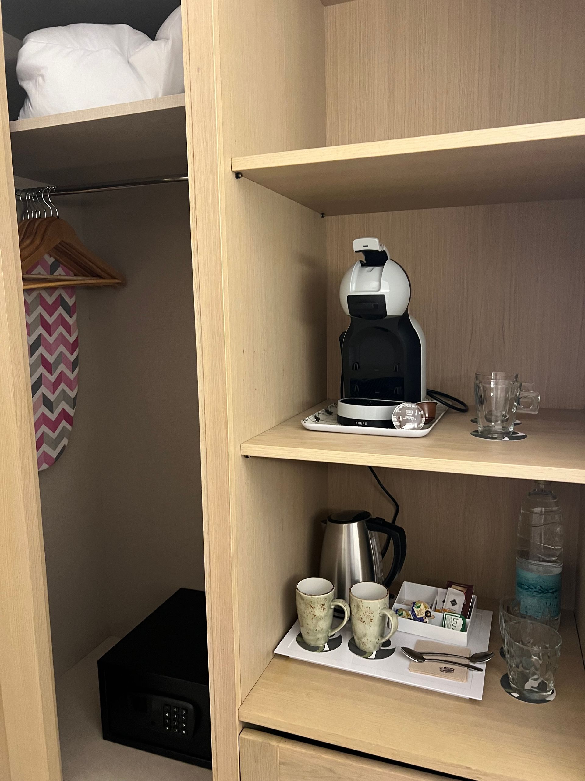 Closet with coffee machine Dolce Gusto, kettle, mugs, glasses, ironing board, safe, and folded towels.