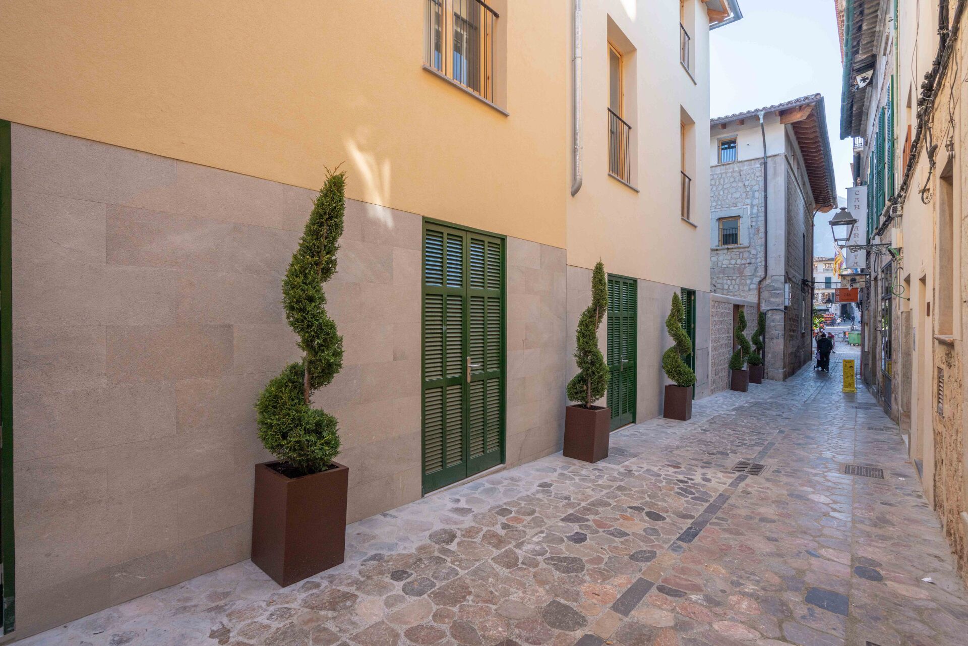 Narrow street in a European town, with trees, green doors, and stone buildings. Street entry to the accessible room.