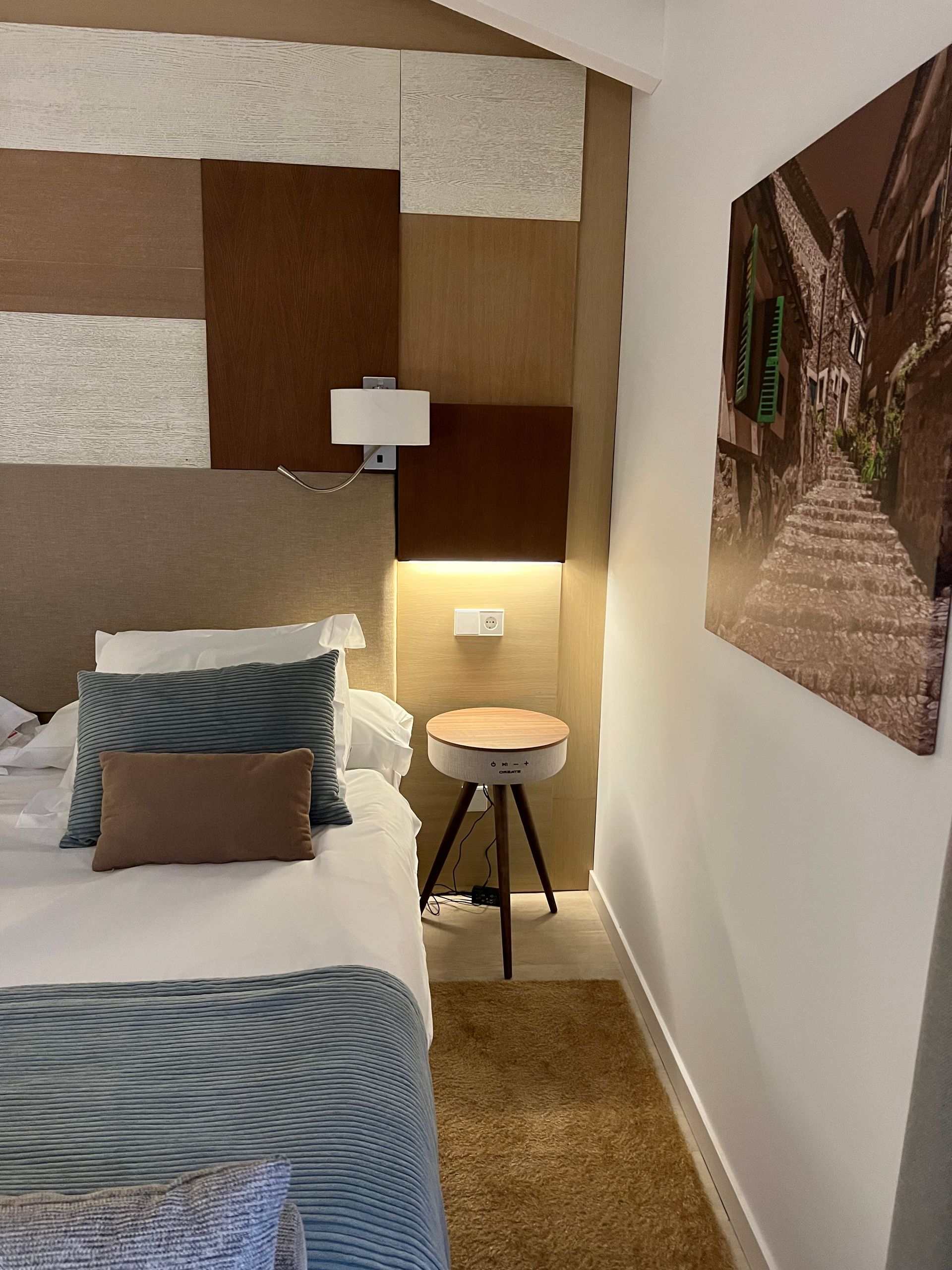 Bedroom corner with bed, side table is also a speaker, artwork, and textured wall panels.