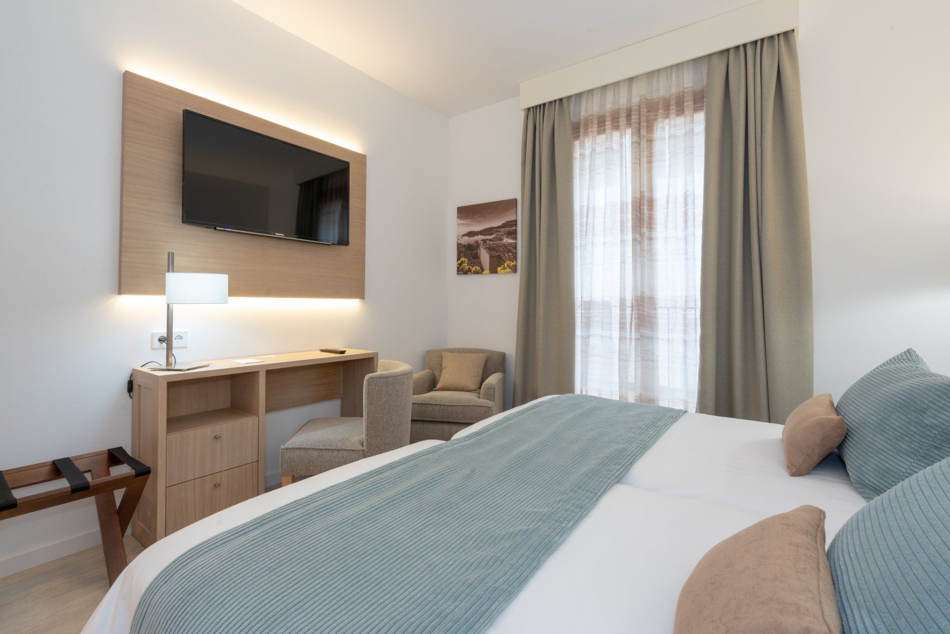 Hotel room with a bed, desk, TV, armchair, and window with curtains.