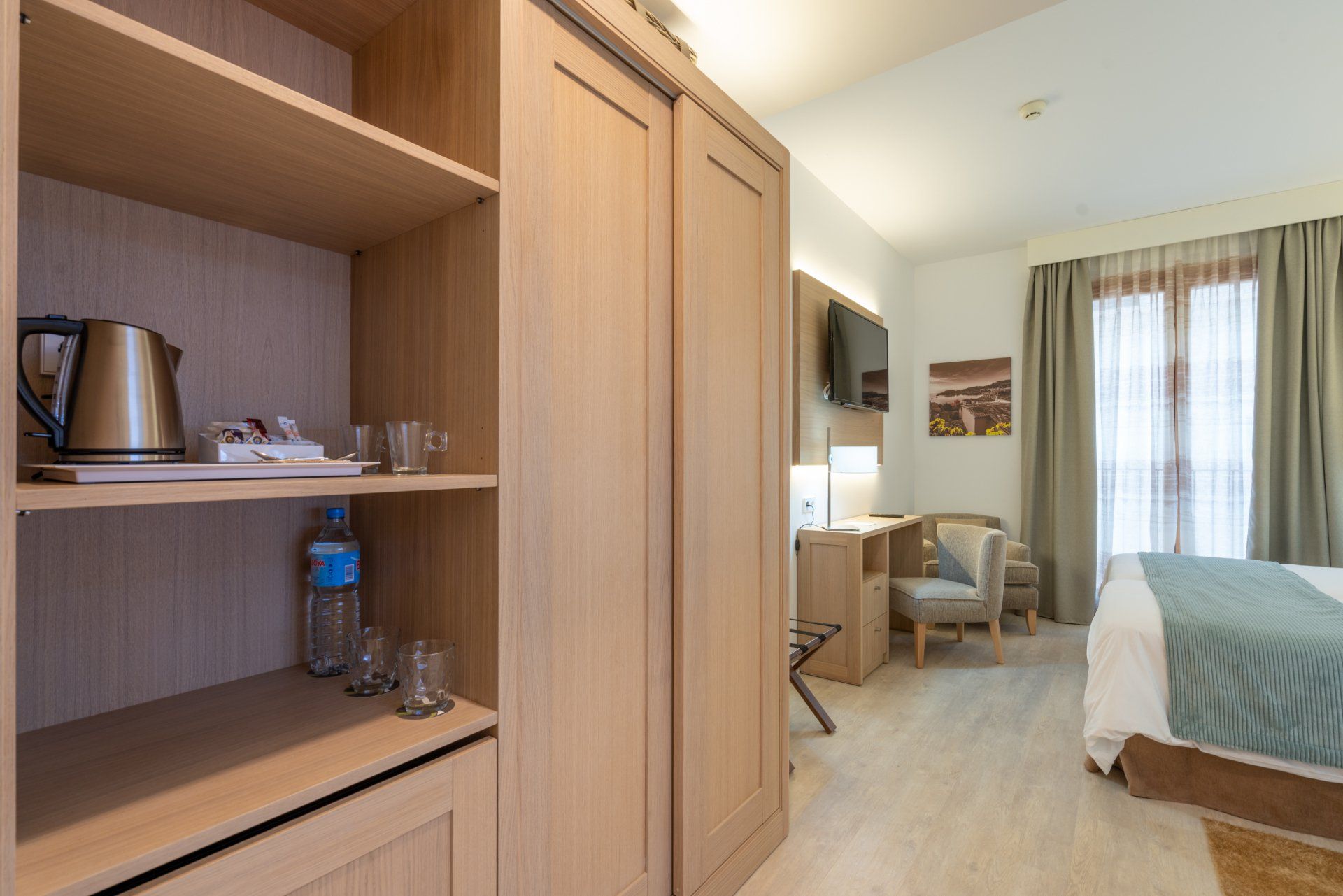 A hotel room with light wood furniture, a desk, bed, and a water kettle.