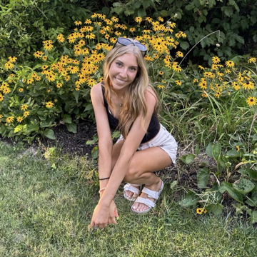 Young caucasian woman bent down next to yellow flowers in white shorts, black tank top, and white sandals. Blonde long hair.