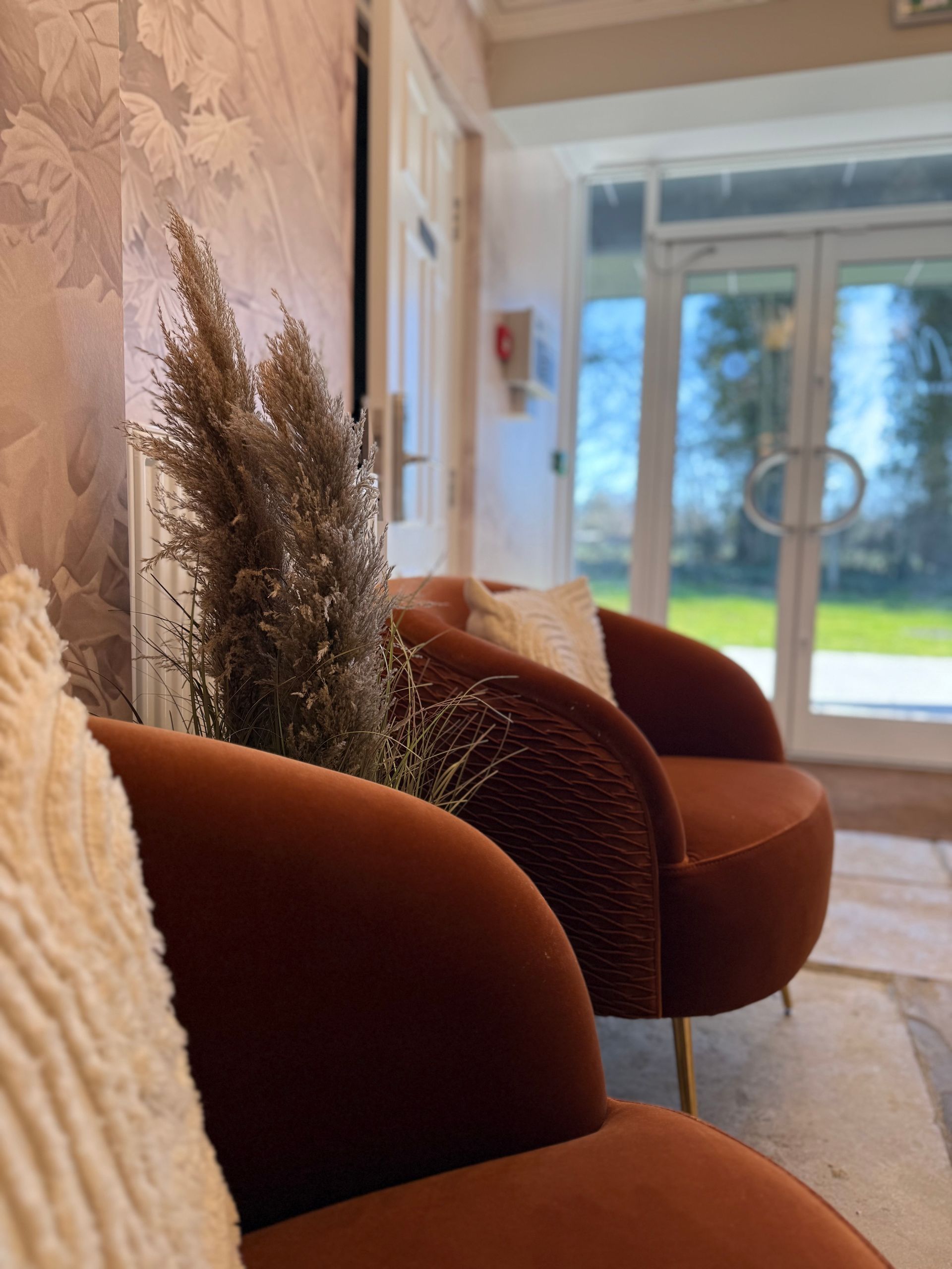 Two orange velvet chairs with a dried floral arrangement; glass doors and light pink wallpaper in the background.