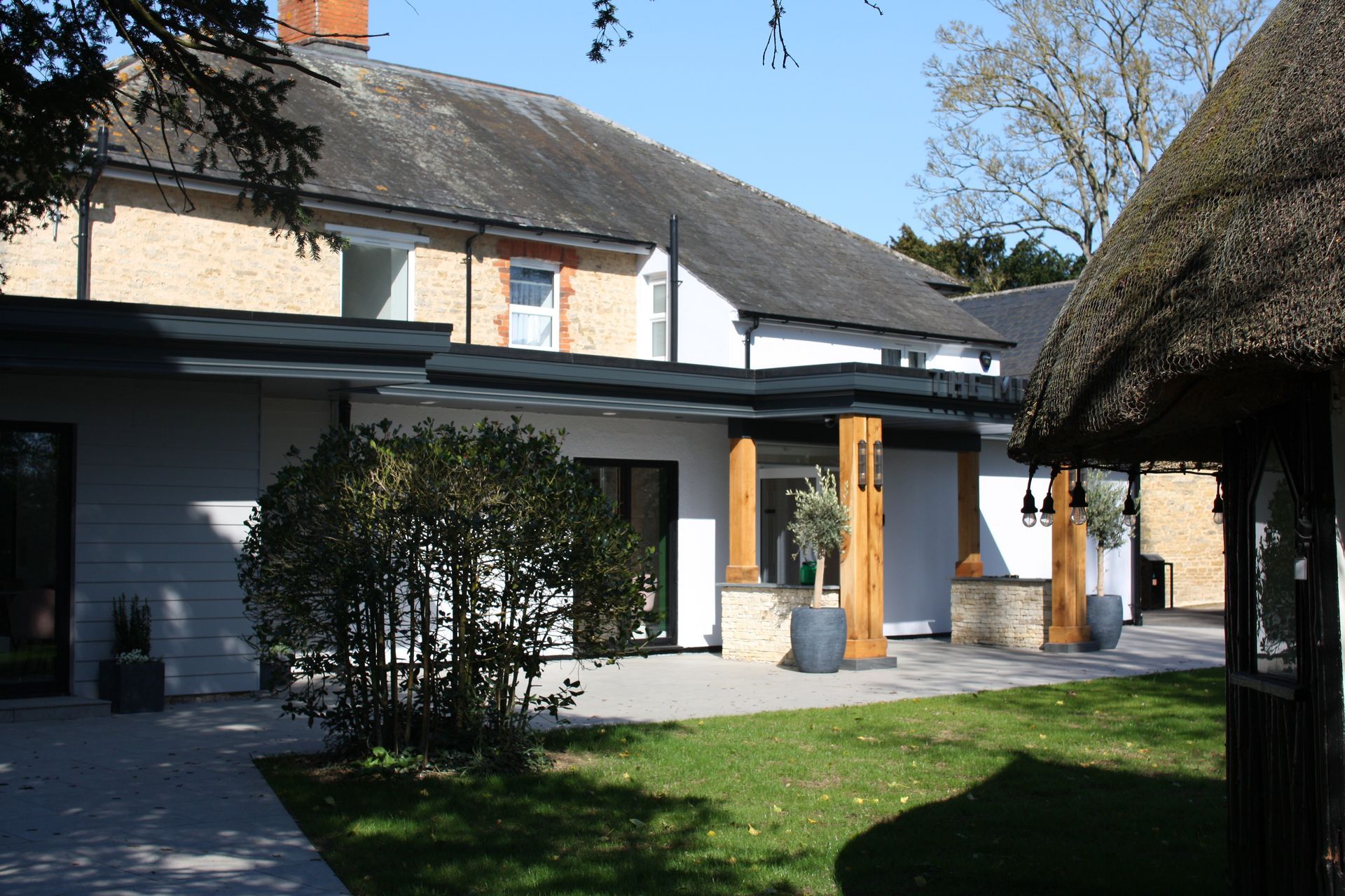White building with stone columns and thatched roof gazebo in a sunny garden.