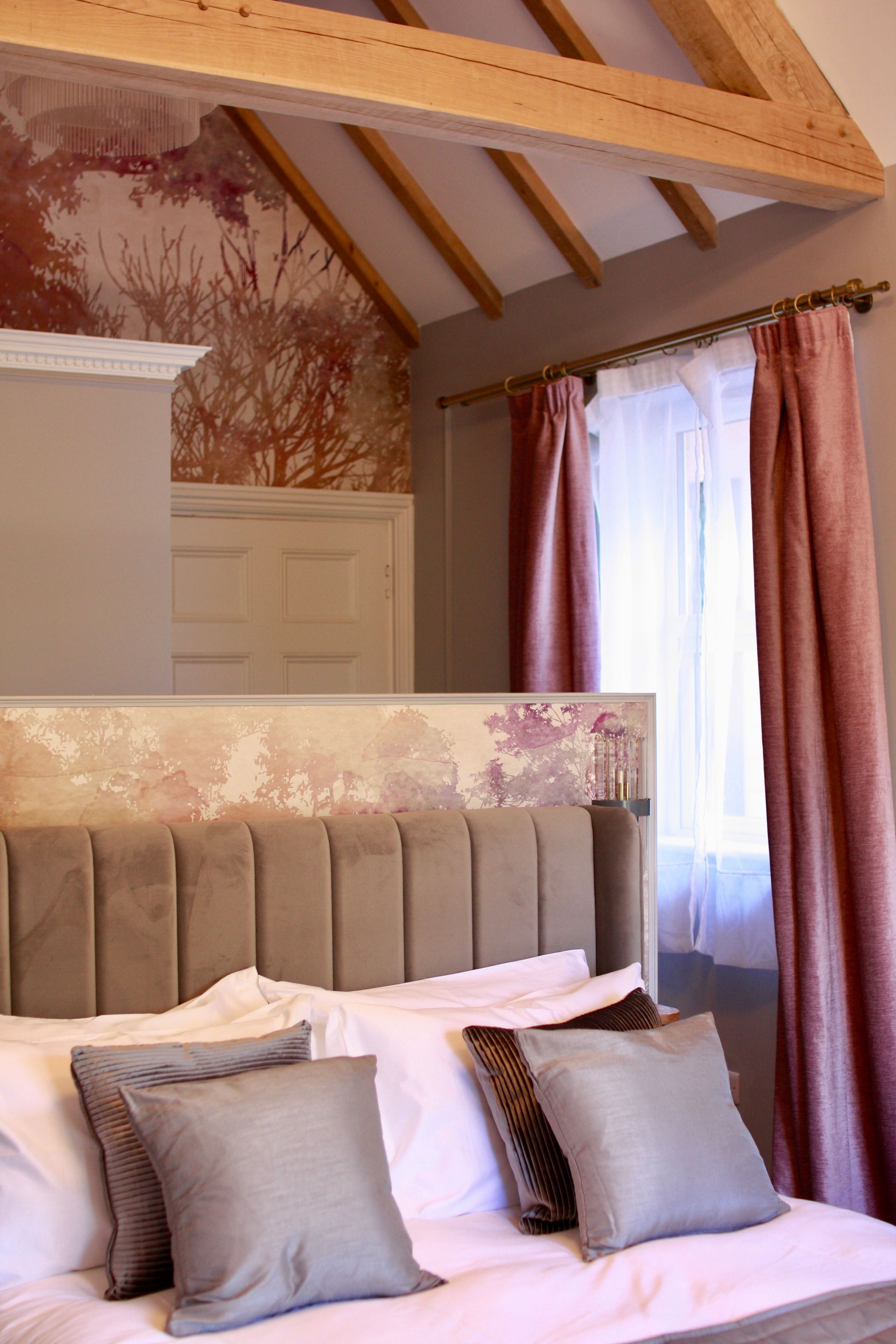 Bedroom with bed, pillows, pink curtains, wood beams, and textured wallpaper.