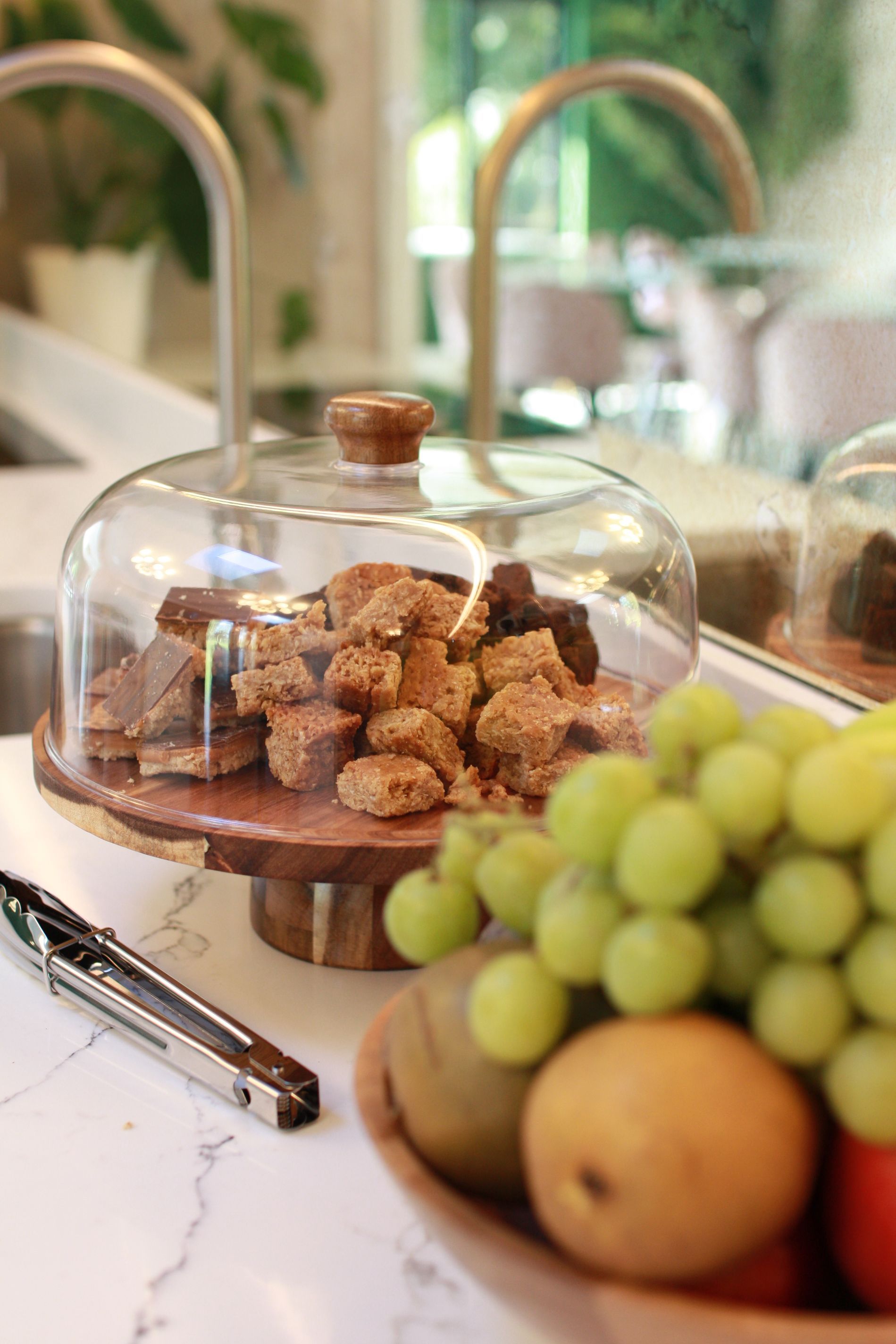 Dessert display with brownies under glass dome, fruit bowl with grapes, kiwi, pear, on marble counter.