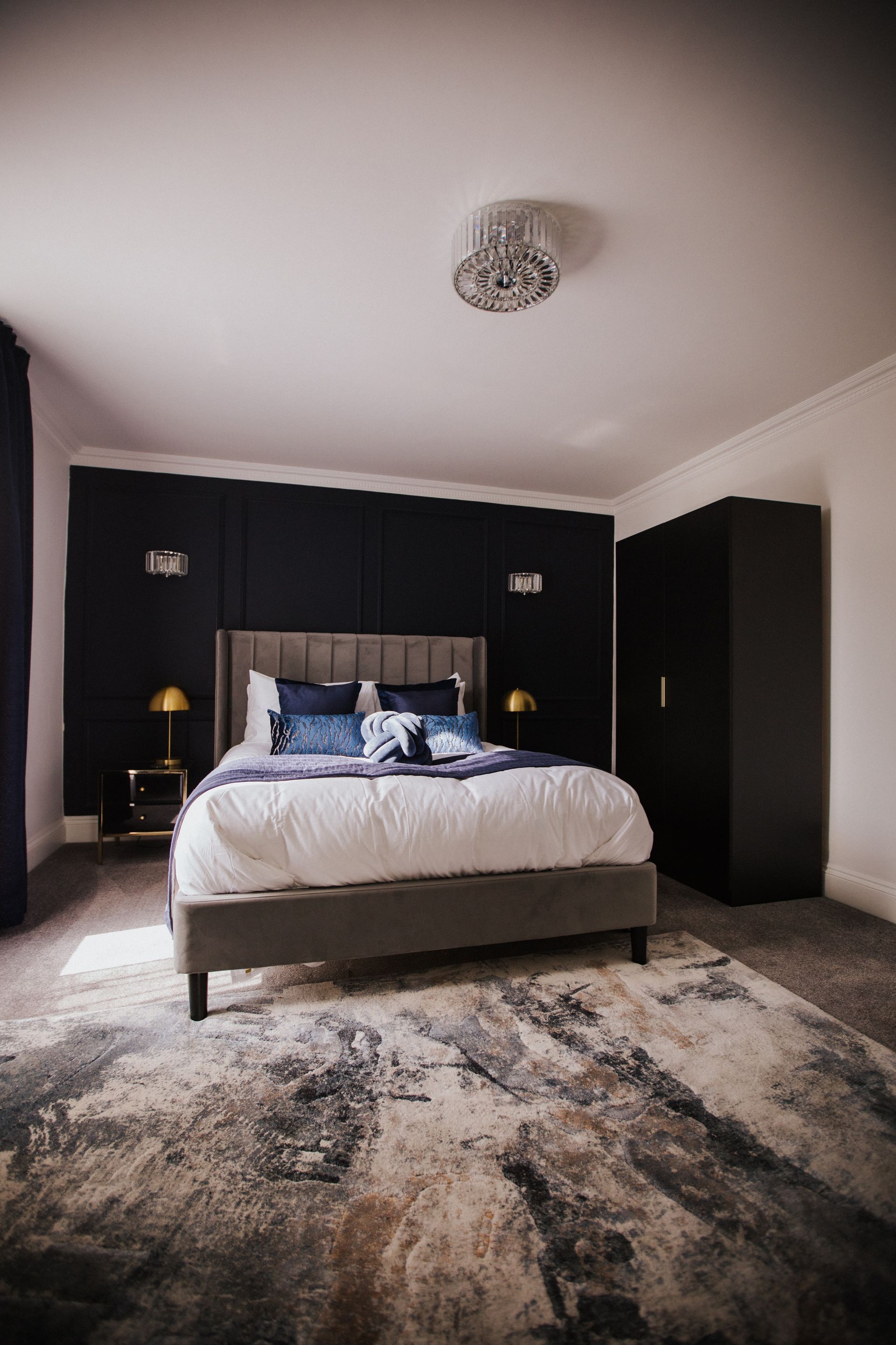 Bedroom interior with dark panel wall, bed, dresser, curtains, and gold chandelier.