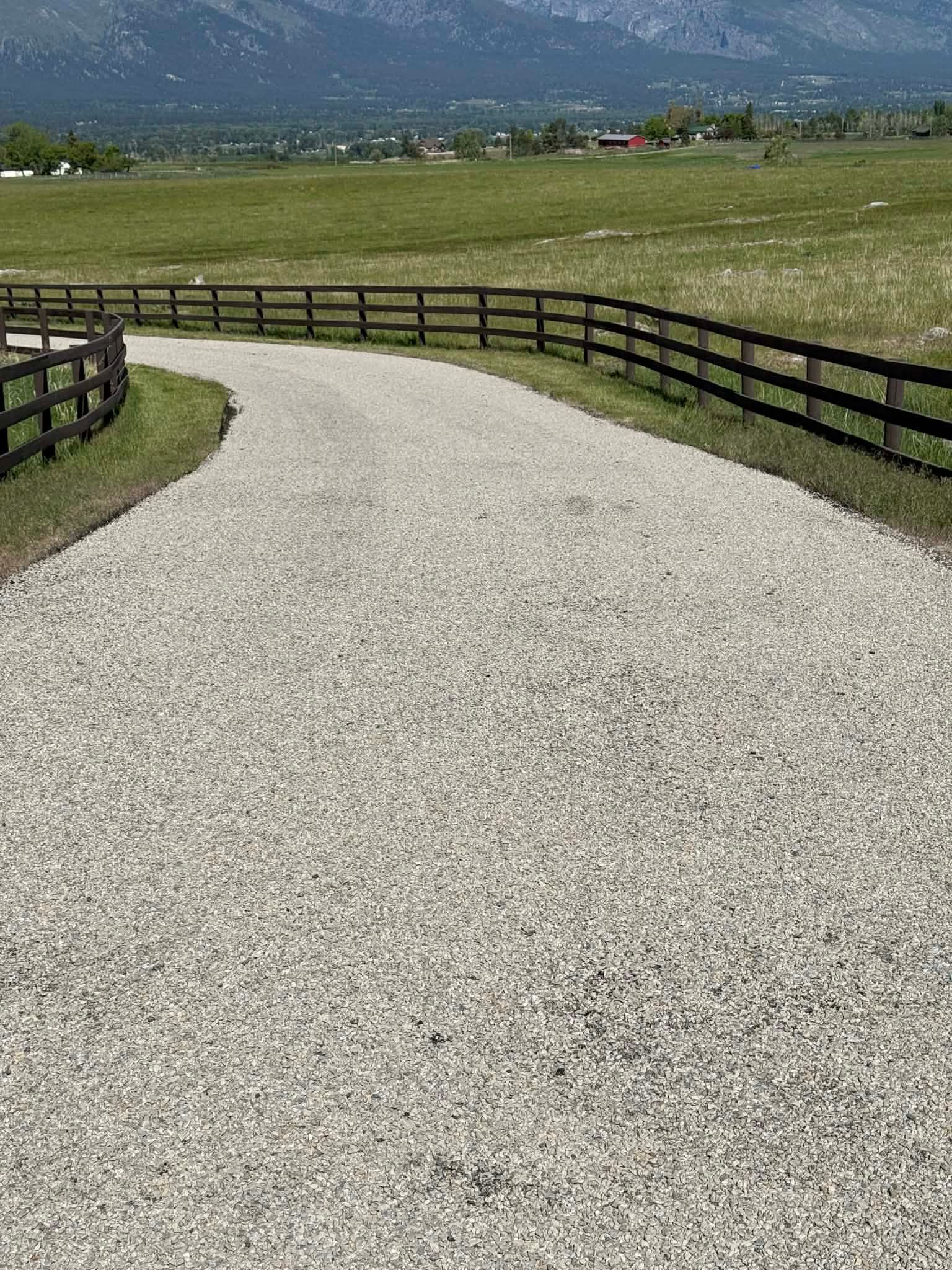 Curving gravel path with wooden fences leading through a grassy field toward distant mountains
