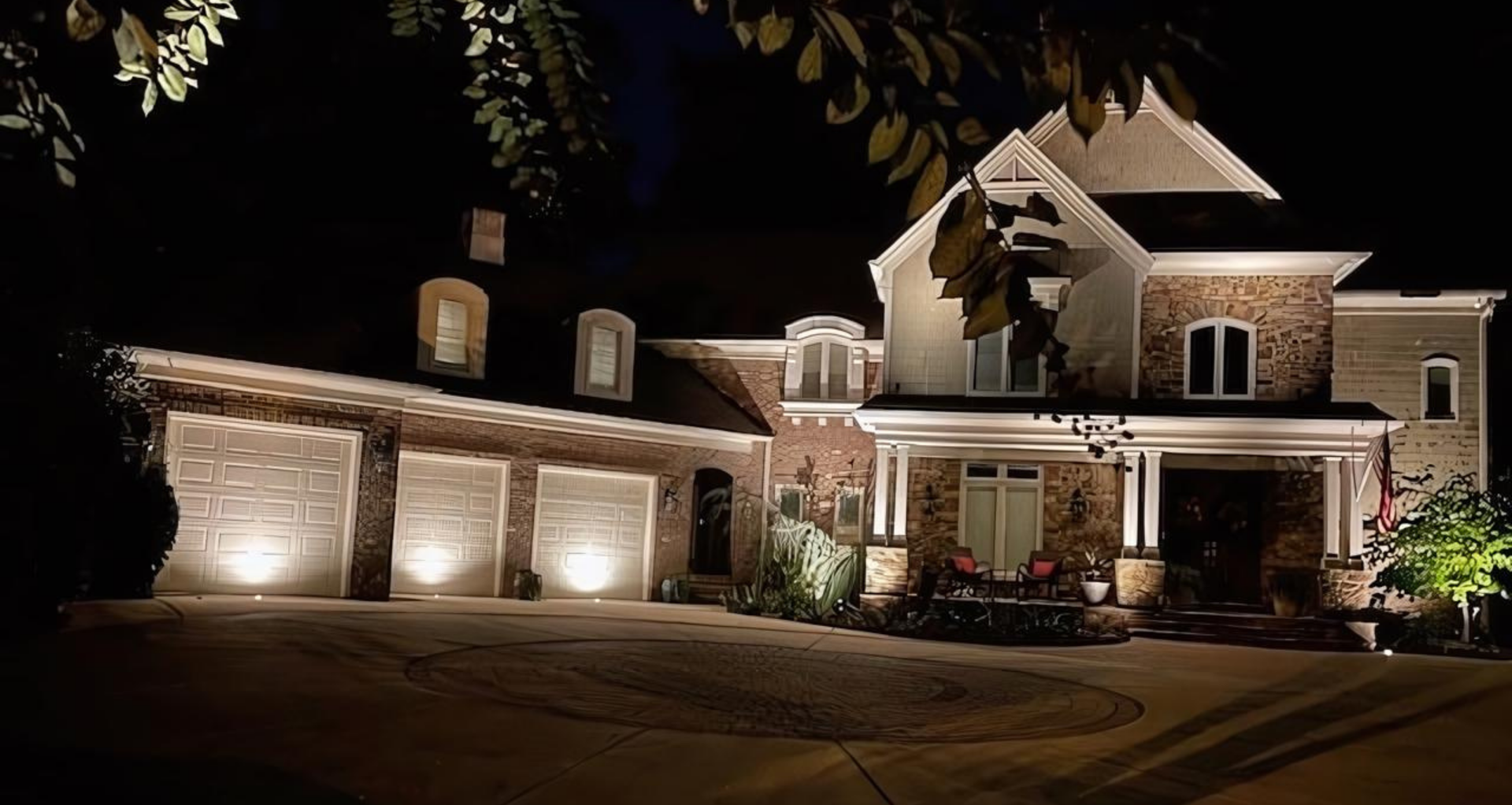 Lit-up luxury house at night. Beige stone and white stucco exterior with three-car garage and entry porch.