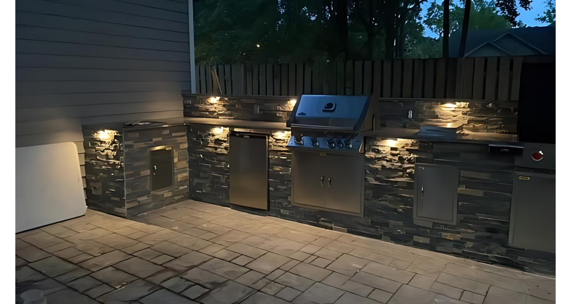 Outdoor kitchen with built-in grill, cabinets, and stone facade, lit by spotlights at night.