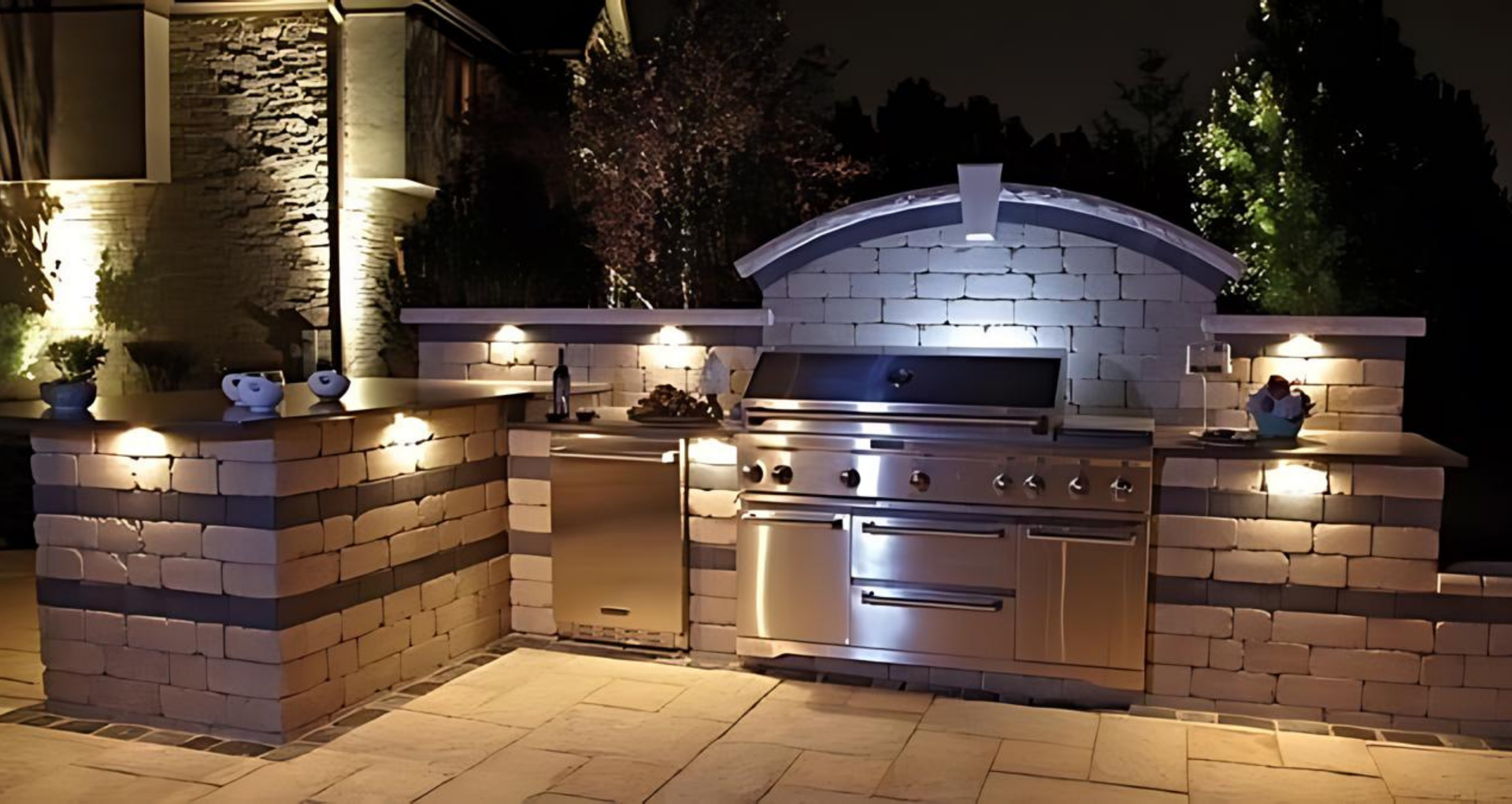 Outdoor kitchen with stainless steel grill, refrigerator, and brick accents, illuminated by night lighting.