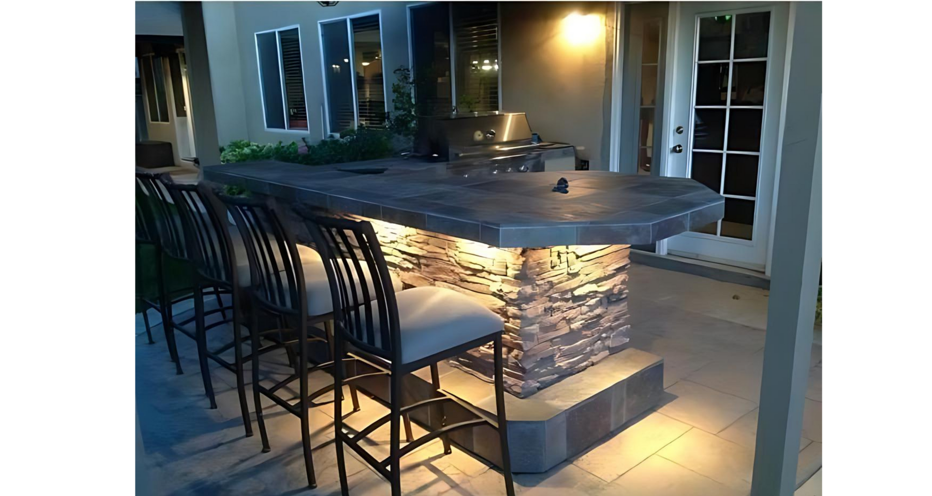 Outdoor bar with stone facade, under-counter lighting, and bar stools. Grill visible. Evening setting.