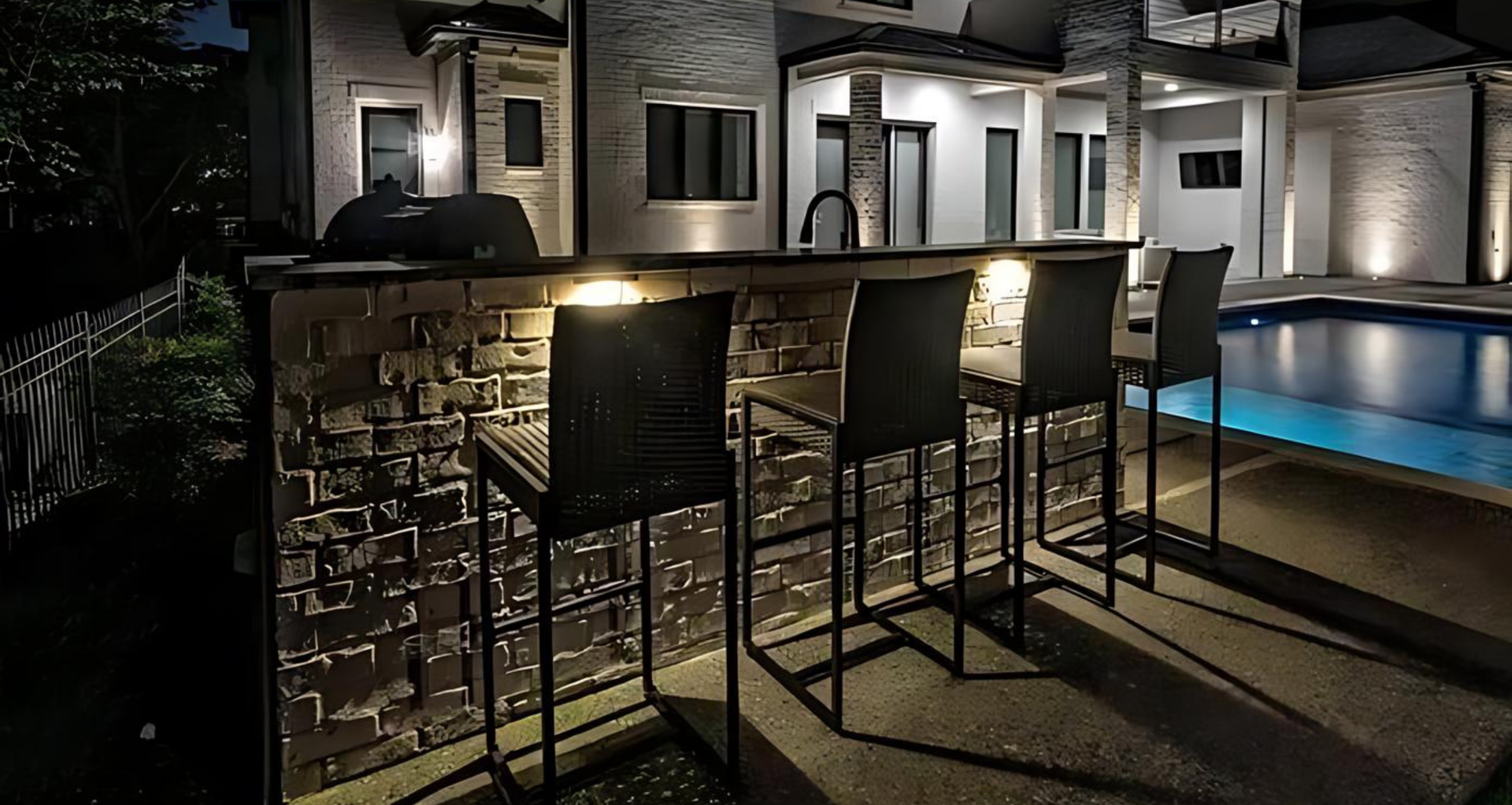 Outdoor bar with four chairs, lit by recessed lights at night, next to a pool.