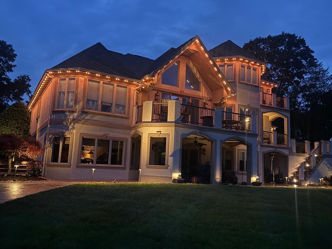 Large, illuminated multi-story house at dusk. Soft lights highlight the architecture. Green lawn in the foreground.