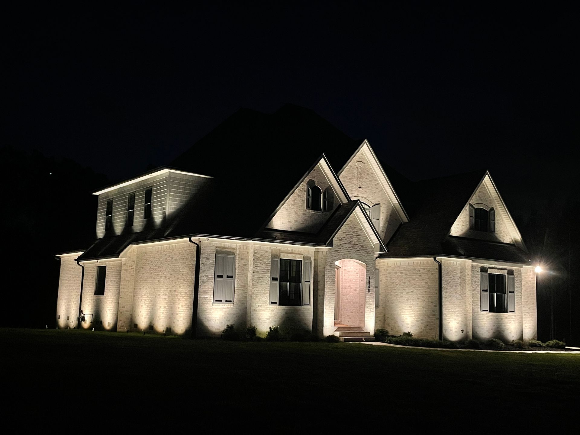 Night view of a brick house illuminated by outdoor lighting, showing the architecture and front lawn.