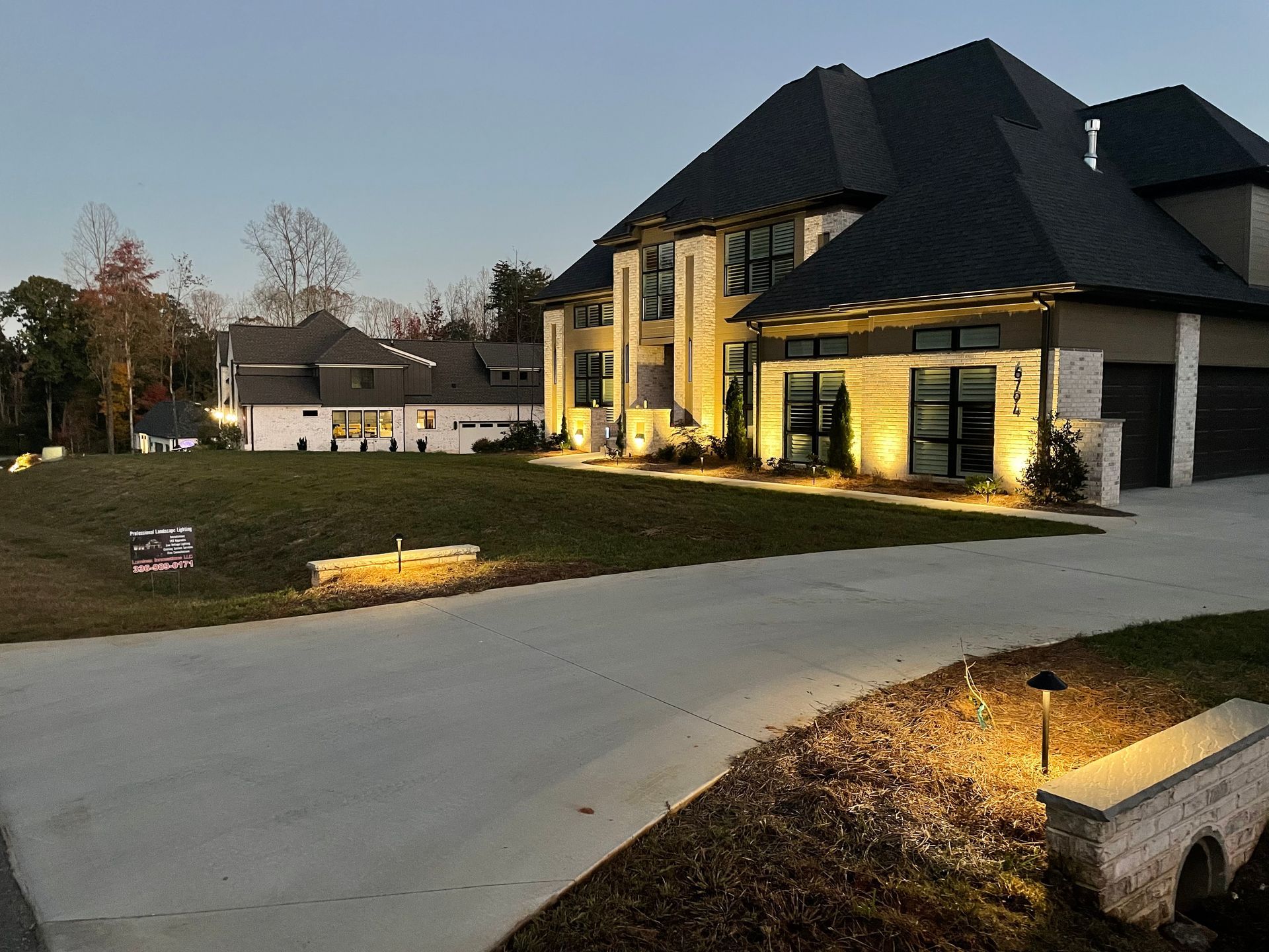Large modern house with exterior lighting at dusk, long driveway, and a neighboring house in the distance.