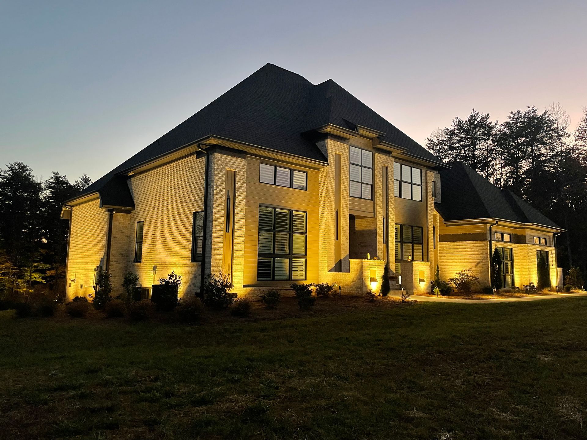 Lit-up two-story brick house at dusk, with several illuminated windows and a dark roof.