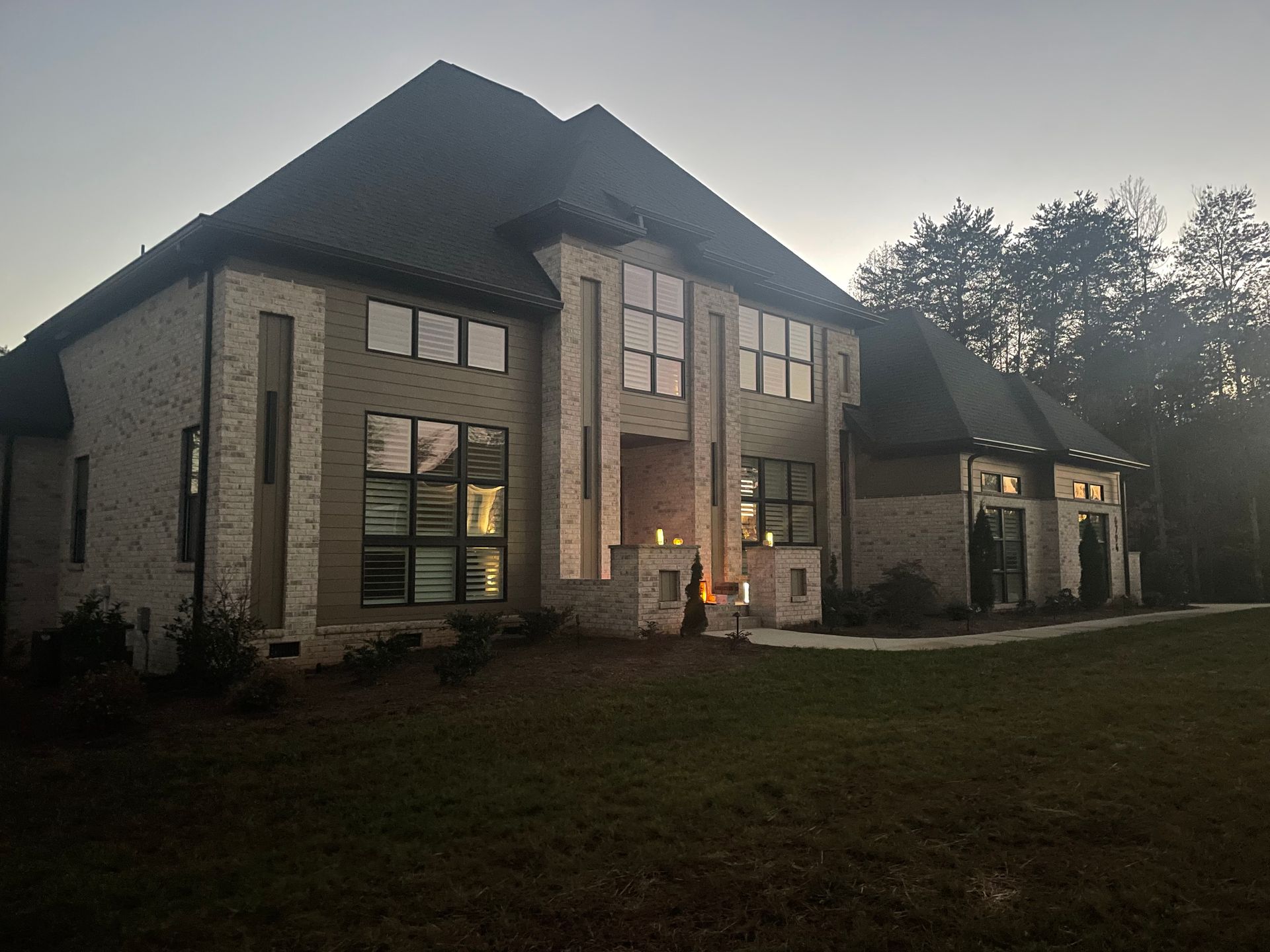 Large, two-story house with light brick and green siding. Large windows reflect the dusk sky.