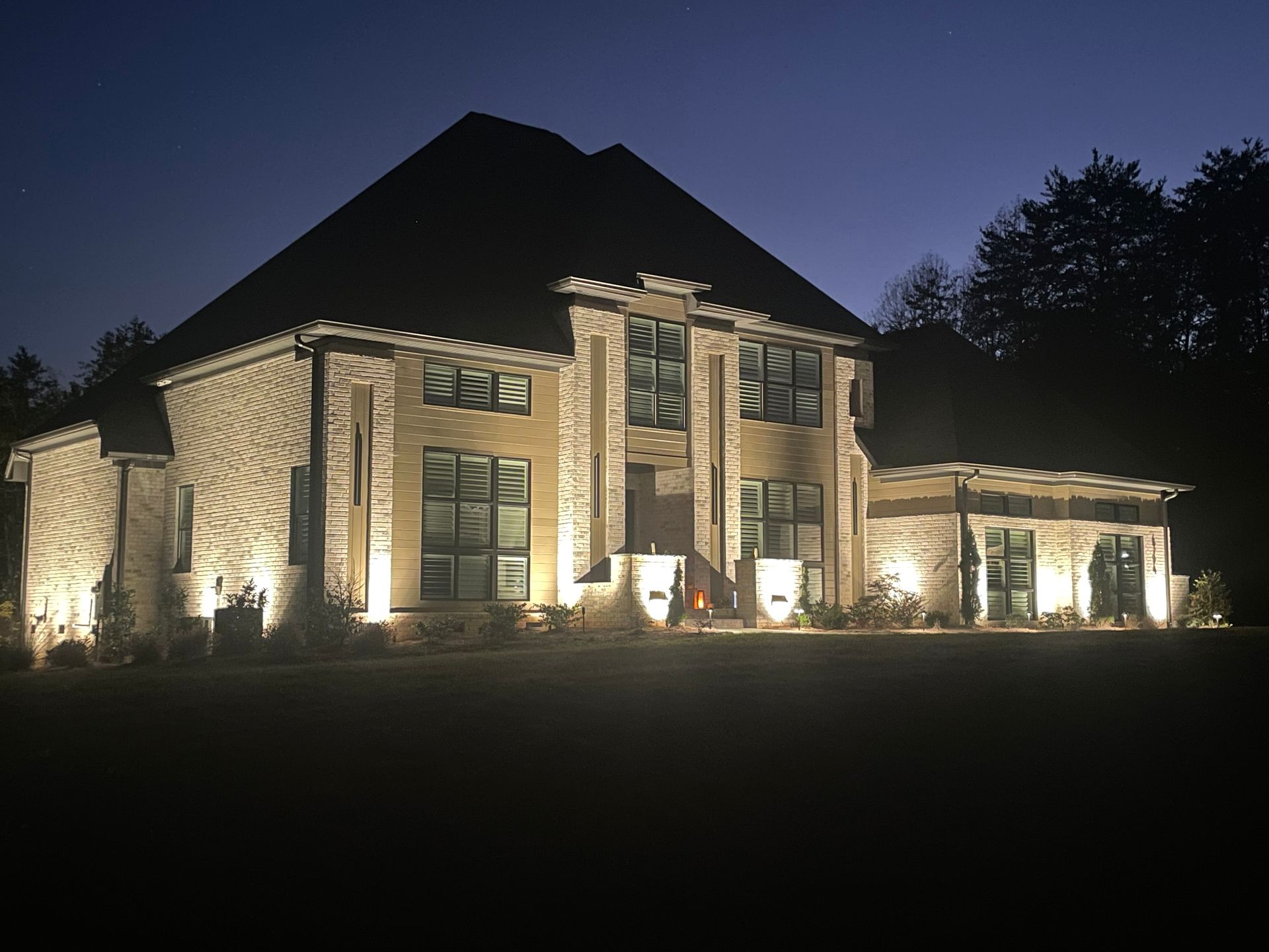 House exterior illuminated at night with landscape lighting. Brick facade, dark roof, windows, and surrounding trees.