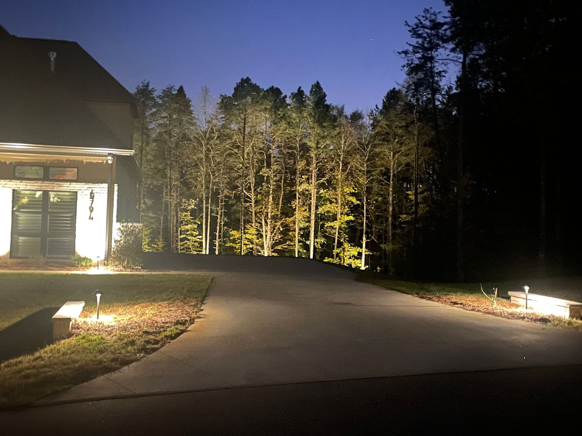 Driveway illuminated by lights leading to a house, with trees lit in the background at dusk.