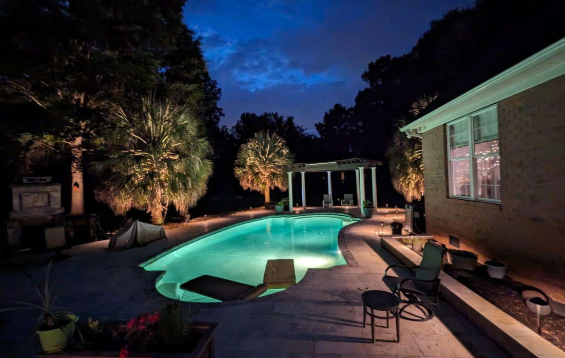 Nighttime backyard with a turquoise pool, illuminated trees, and a house with lit windows.