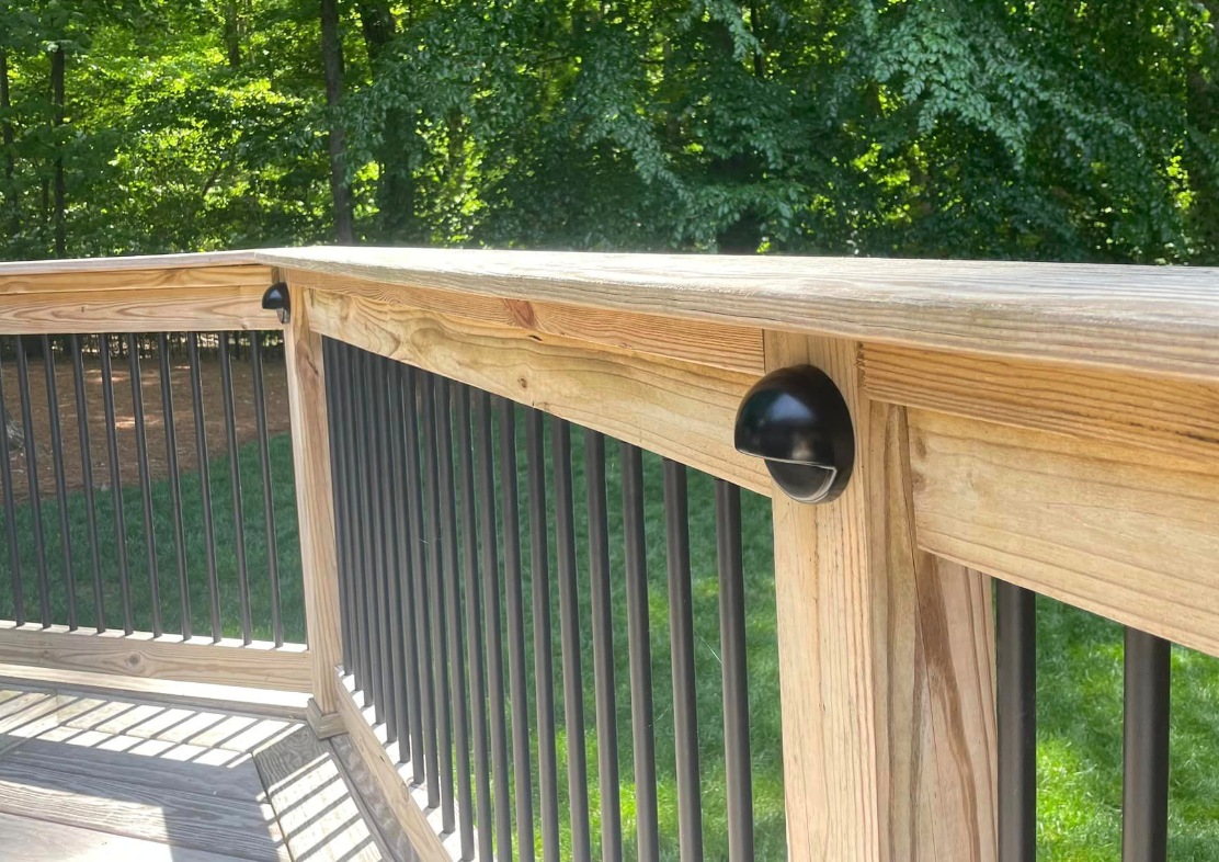 Wooden deck railing with black vertical spindles and black lights, against green trees.
