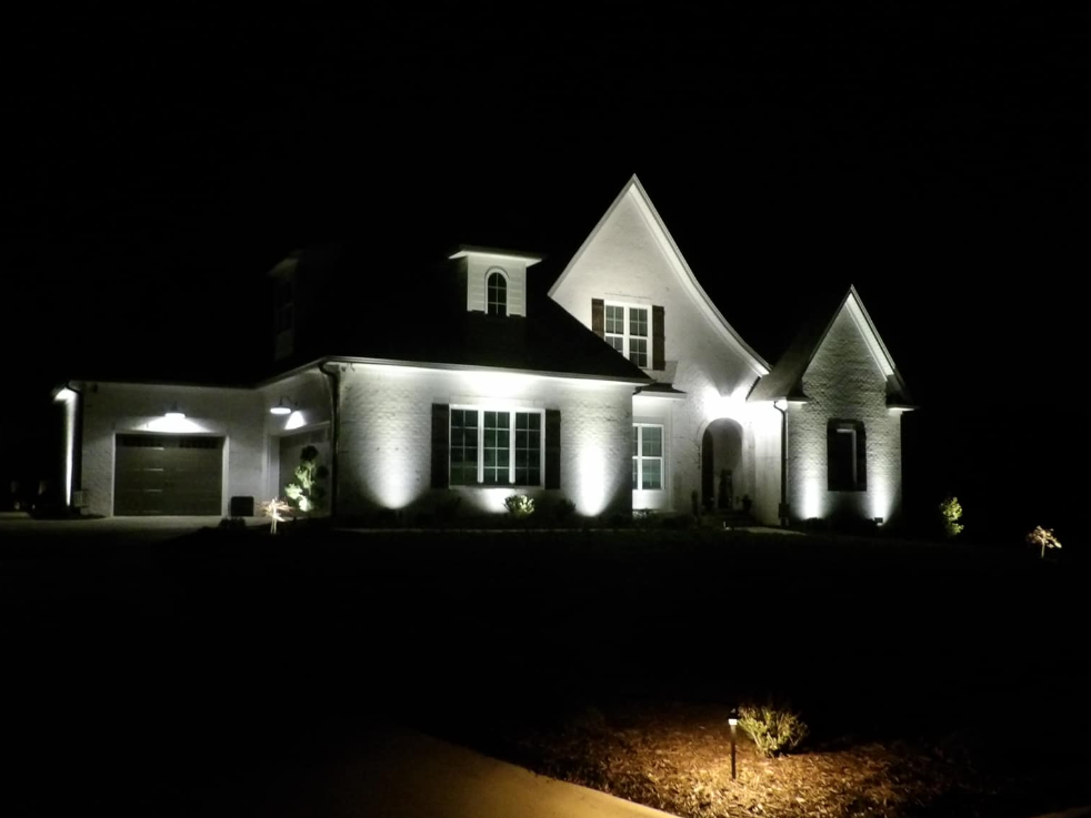 A brightly lit white brick house at night, illuminated by outdoor lights in a dark setting.