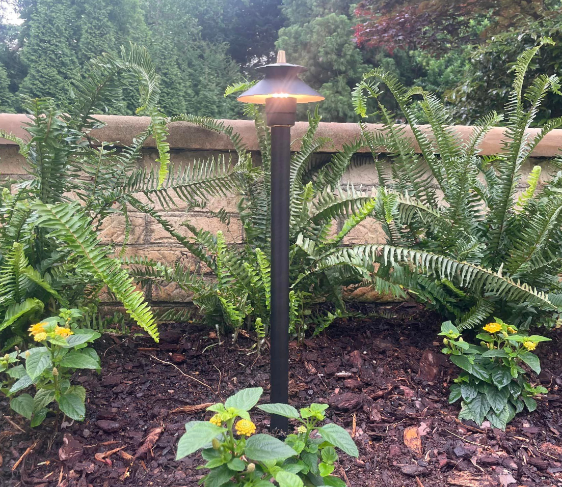 Black outdoor lamp post in a garden bed with lush green plants and a tan brick wall.