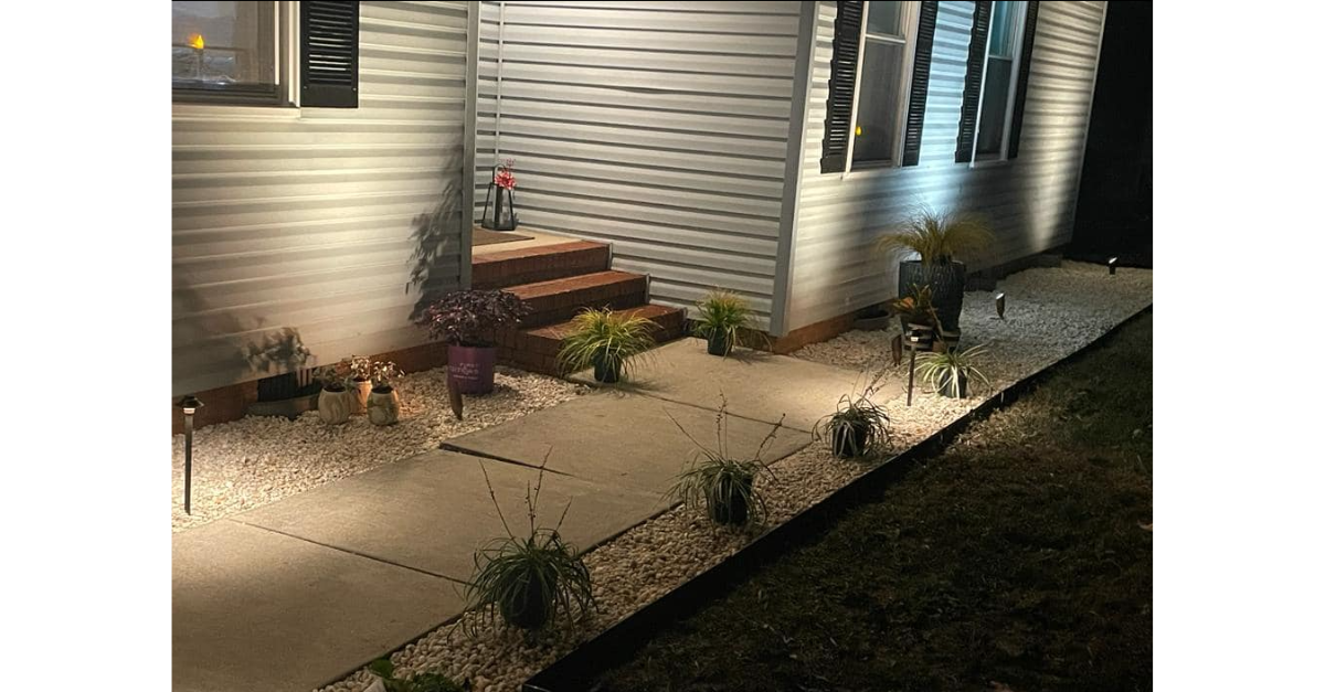Lit walkway and landscaping in front of a house at night, featuring plants, gravel, and exterior lights.