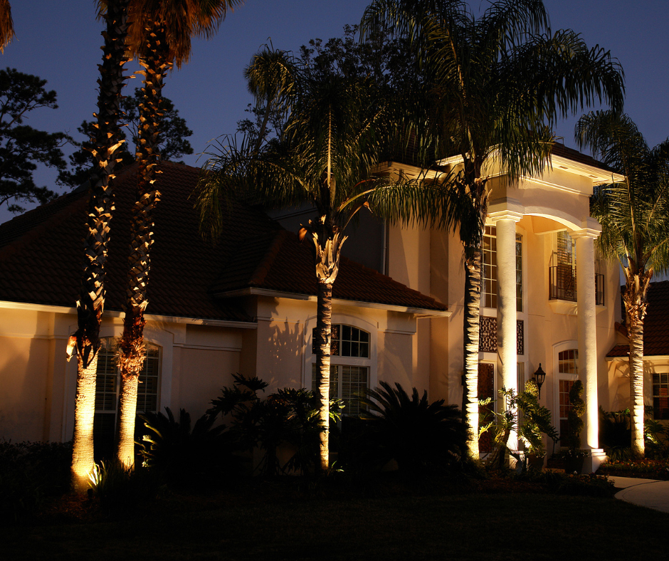 Lit-up luxury house at night, illuminated palm trees in front, with bright columns, and dark sky.