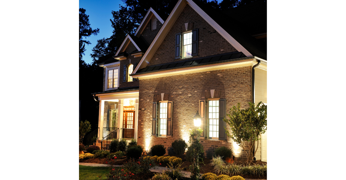 A brick house illuminated at night with garden lighting, bushes, and an open front door.