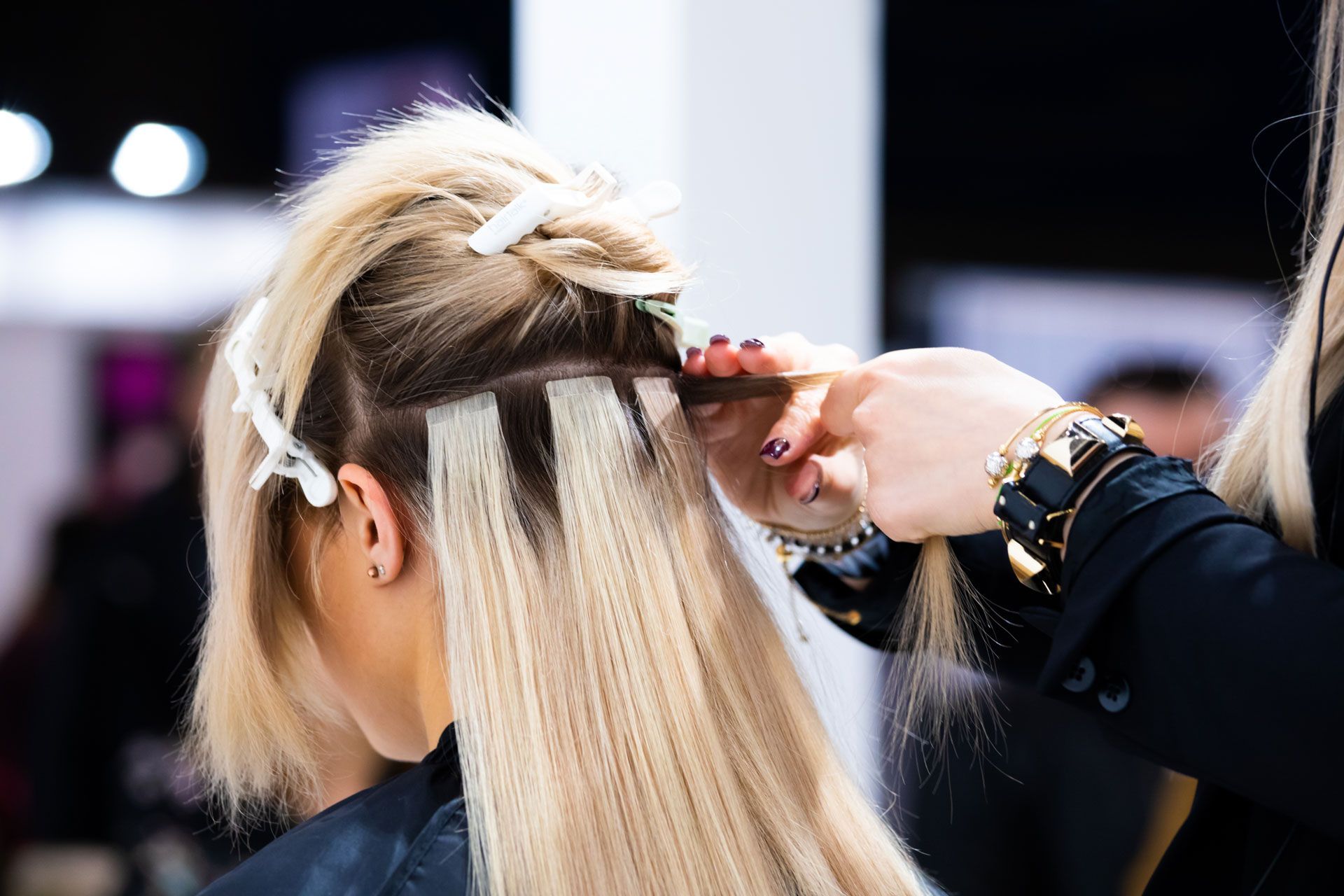 Stylist applying hair extensions to a client with blonde hair, in a salon setting.