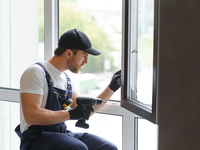 A man is sitting on a window sill using a drill to install a window.
