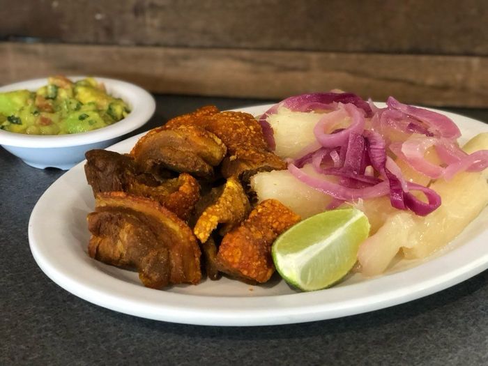 A white plate topped with meat , potatoes and onions next to a bowl of guacamole.