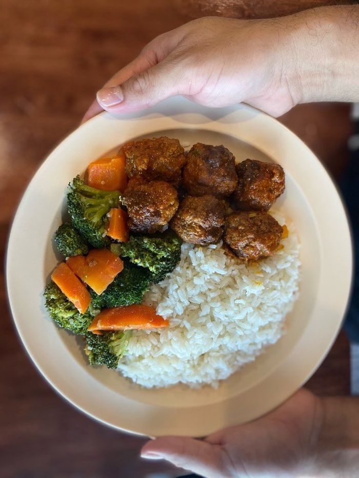 A person is holding a plate of food with meatballs , broccoli , carrots and rice.