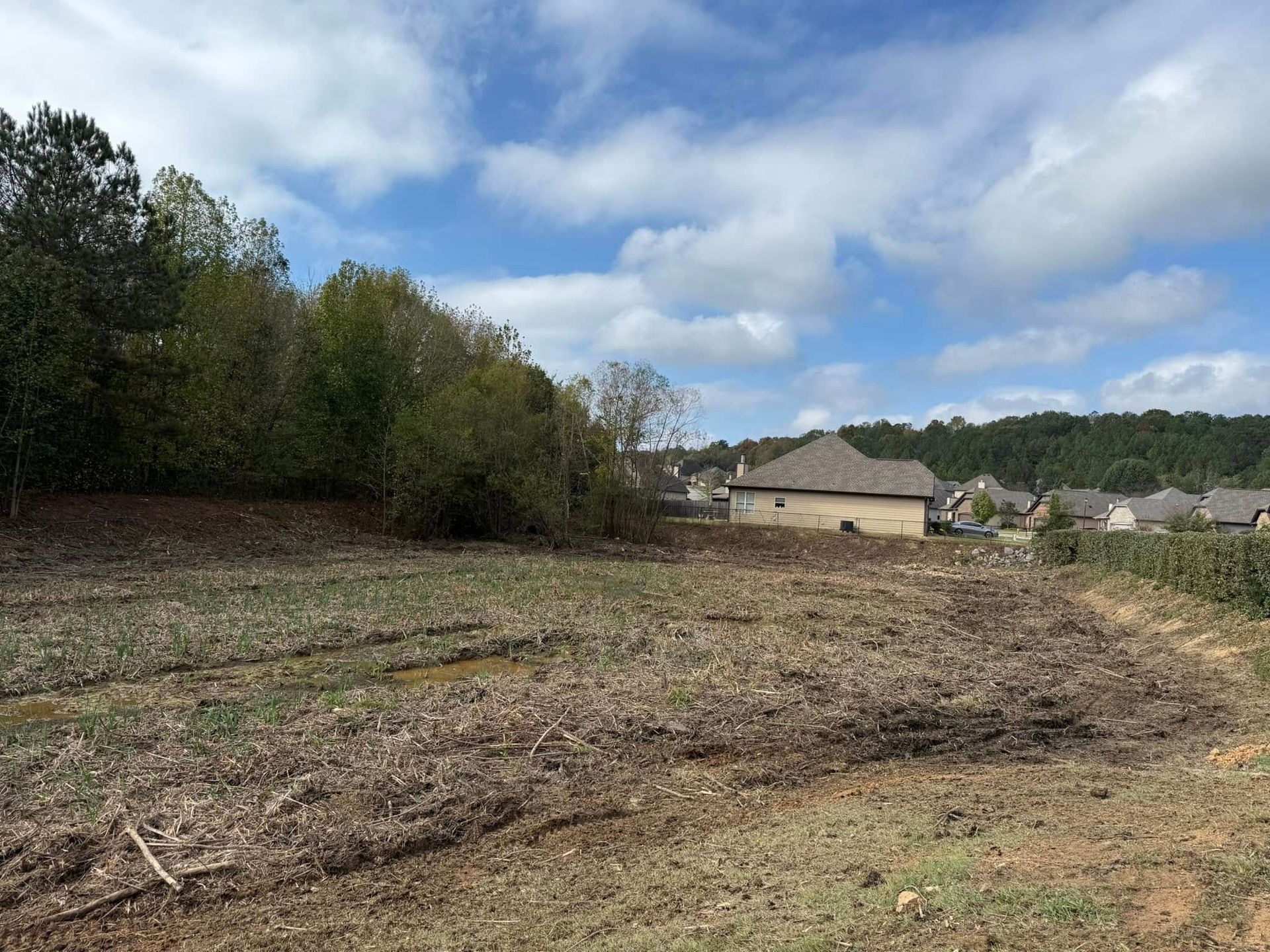 A cleared, muddy lot sits in the foreground with a wooded area to the left and a residential neighborhood in the distance.