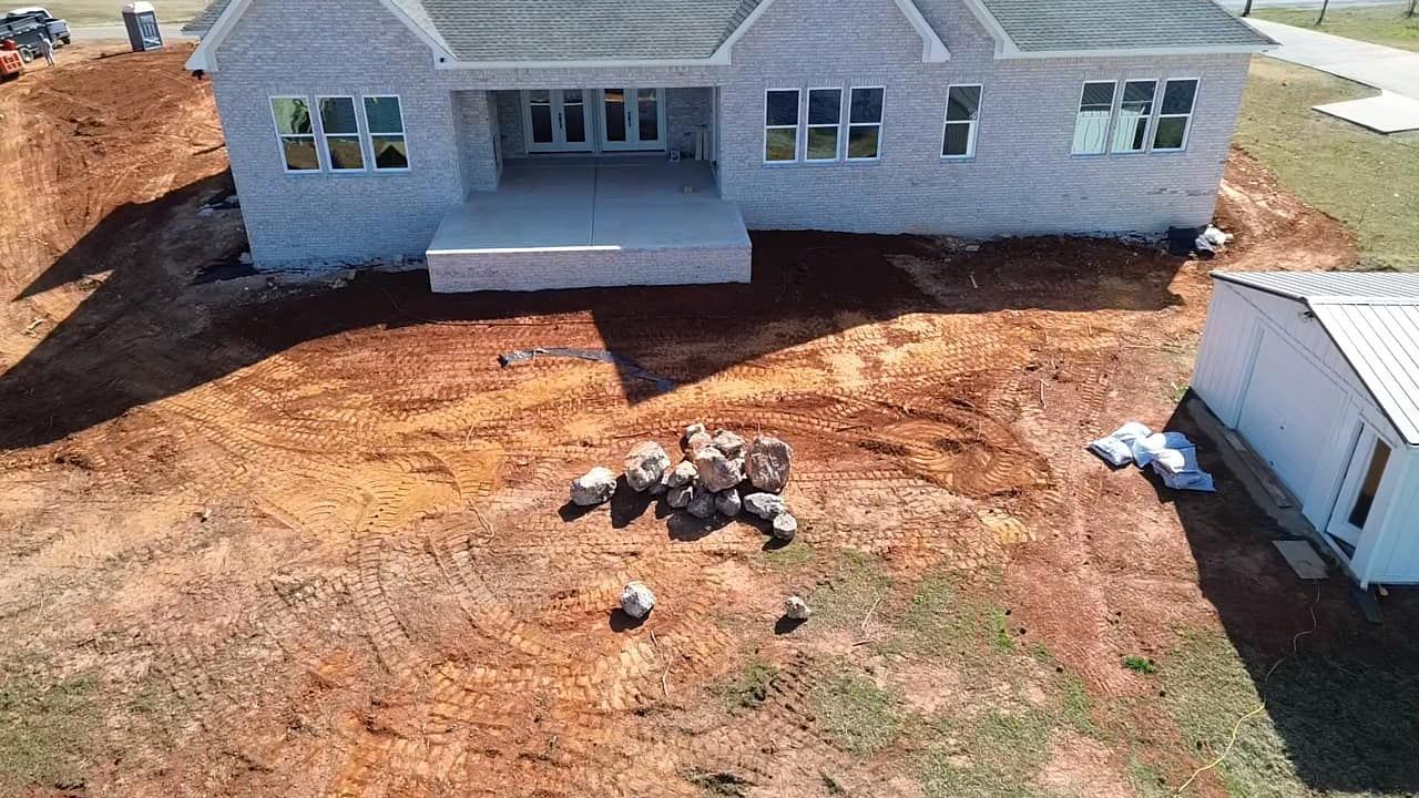 An aerial view of a light-colored brick house under construction, surrounded by reddish-brown dirt and a small outbuilding.