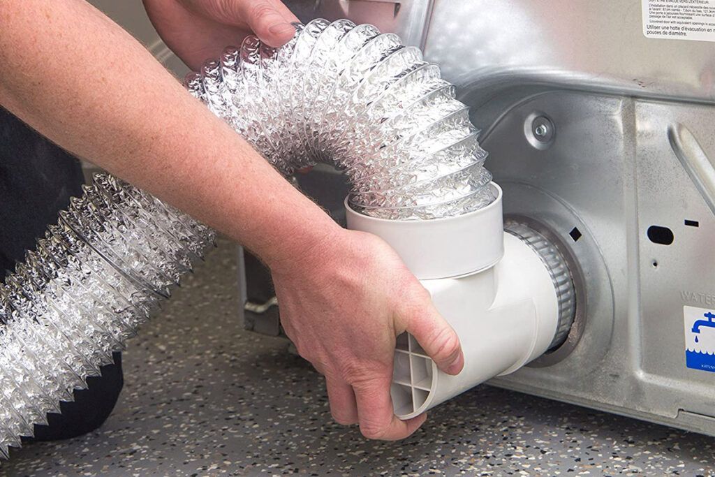 Hands connecting a silver dryer vent hose to a white appliance duct outlet.