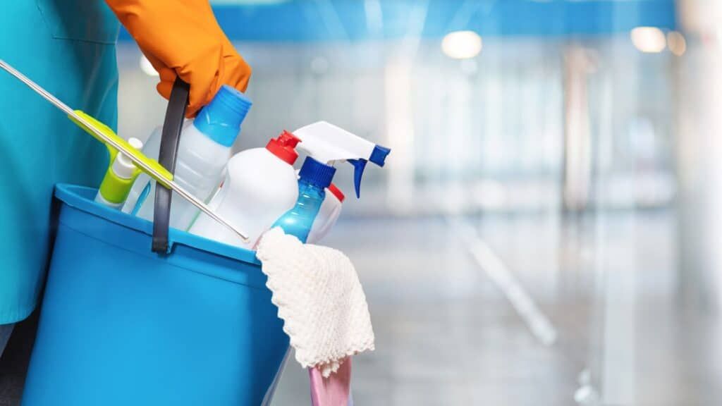 Cleaning supplies in a blue bucket held by gloved hands, with spray bottles and a mop in a bright hallway