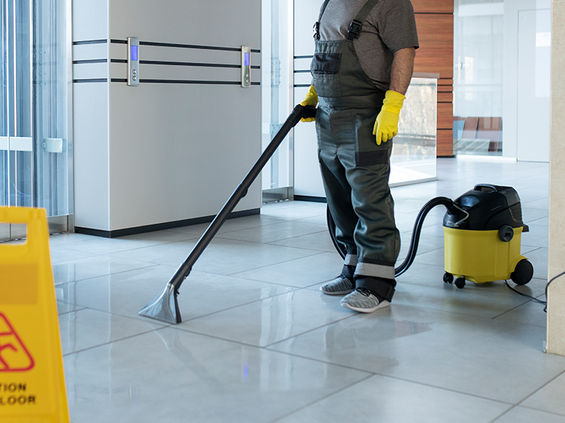 Person vacuuming a shiny office hallway with a yellow canister vacuum and wearing gloves and boots