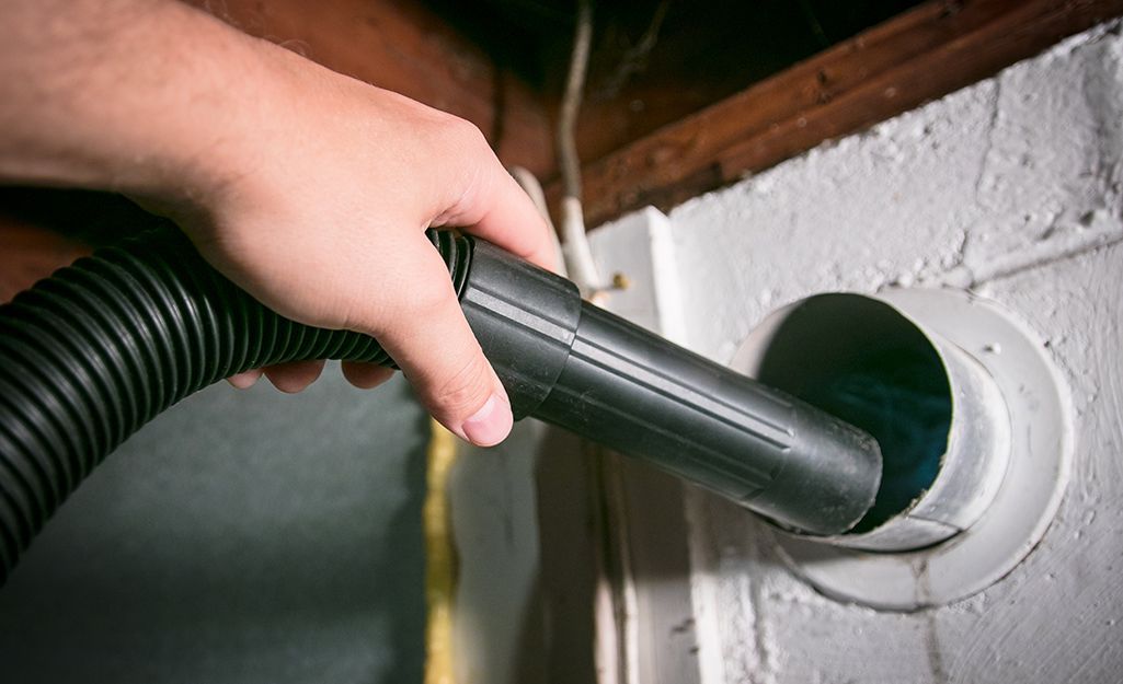 Hand inserting a vacuum hose into a white wall vent in a basement