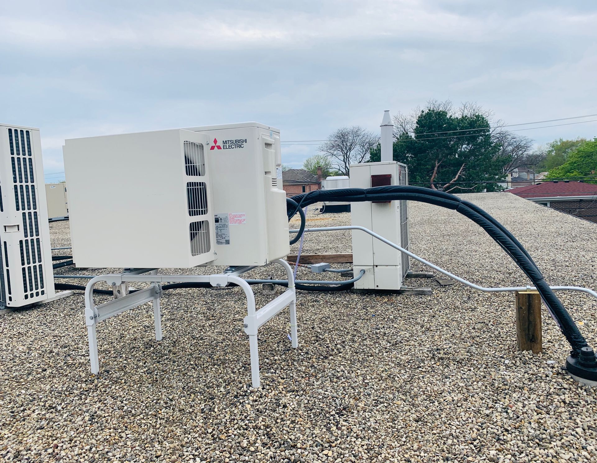 Outdoor HVAC unit with thick black hoses on a gravel rooftop, near a white metal fence and trees