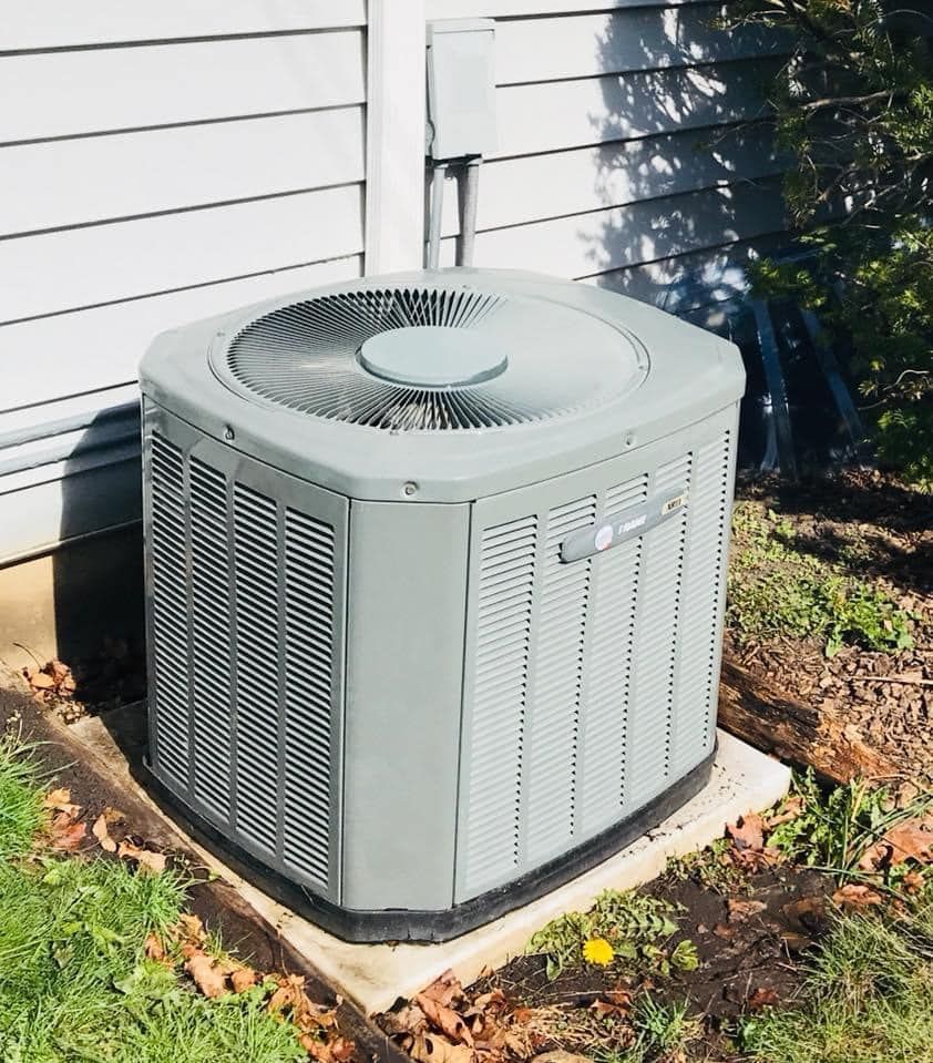 Outdoor central air conditioning unit beside a house, sitting on a concrete pad with grass and leaves nearby