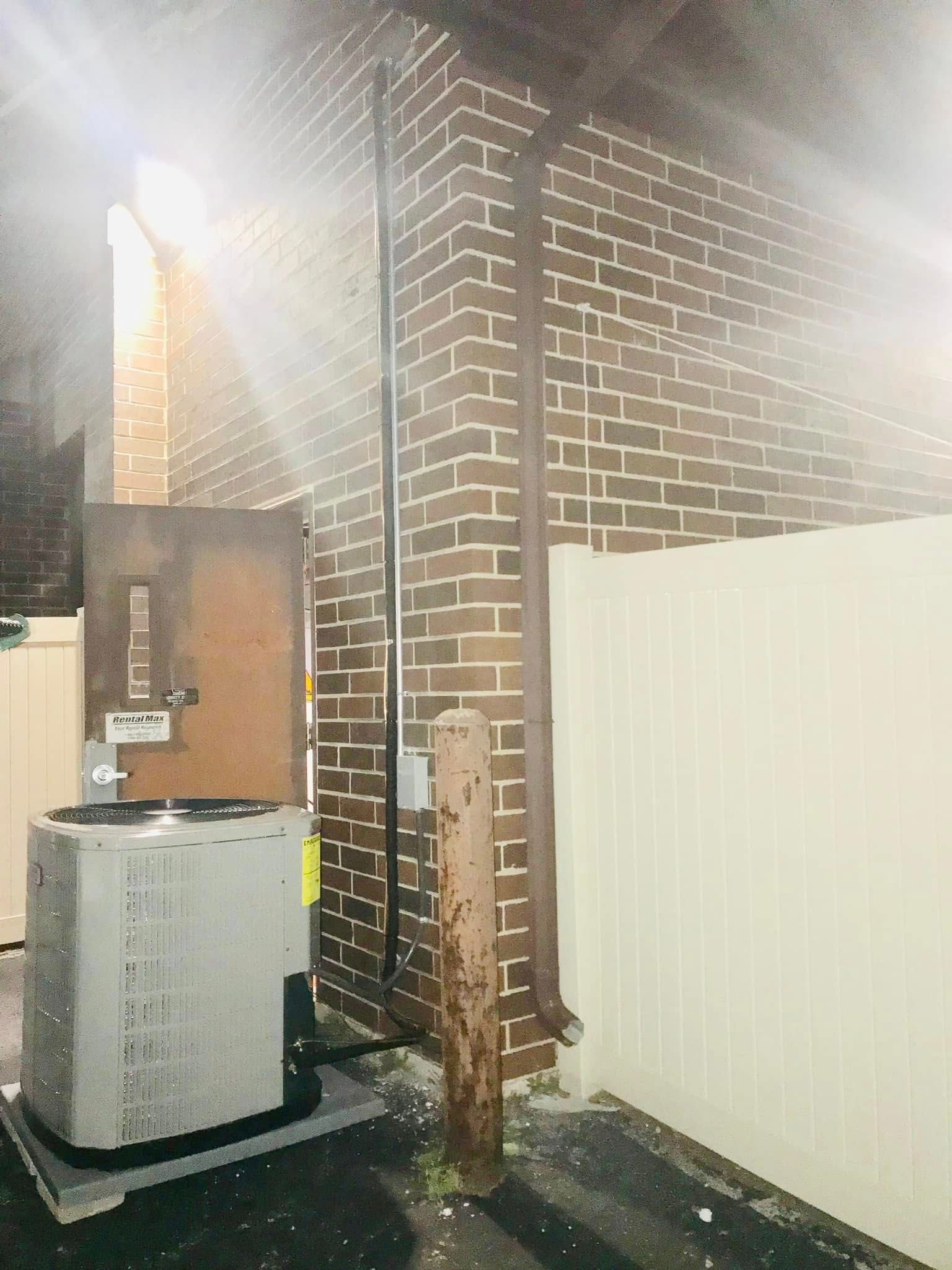 Outdoor HVAC unit beside a brick house and white fence at night, lit by a bright light.