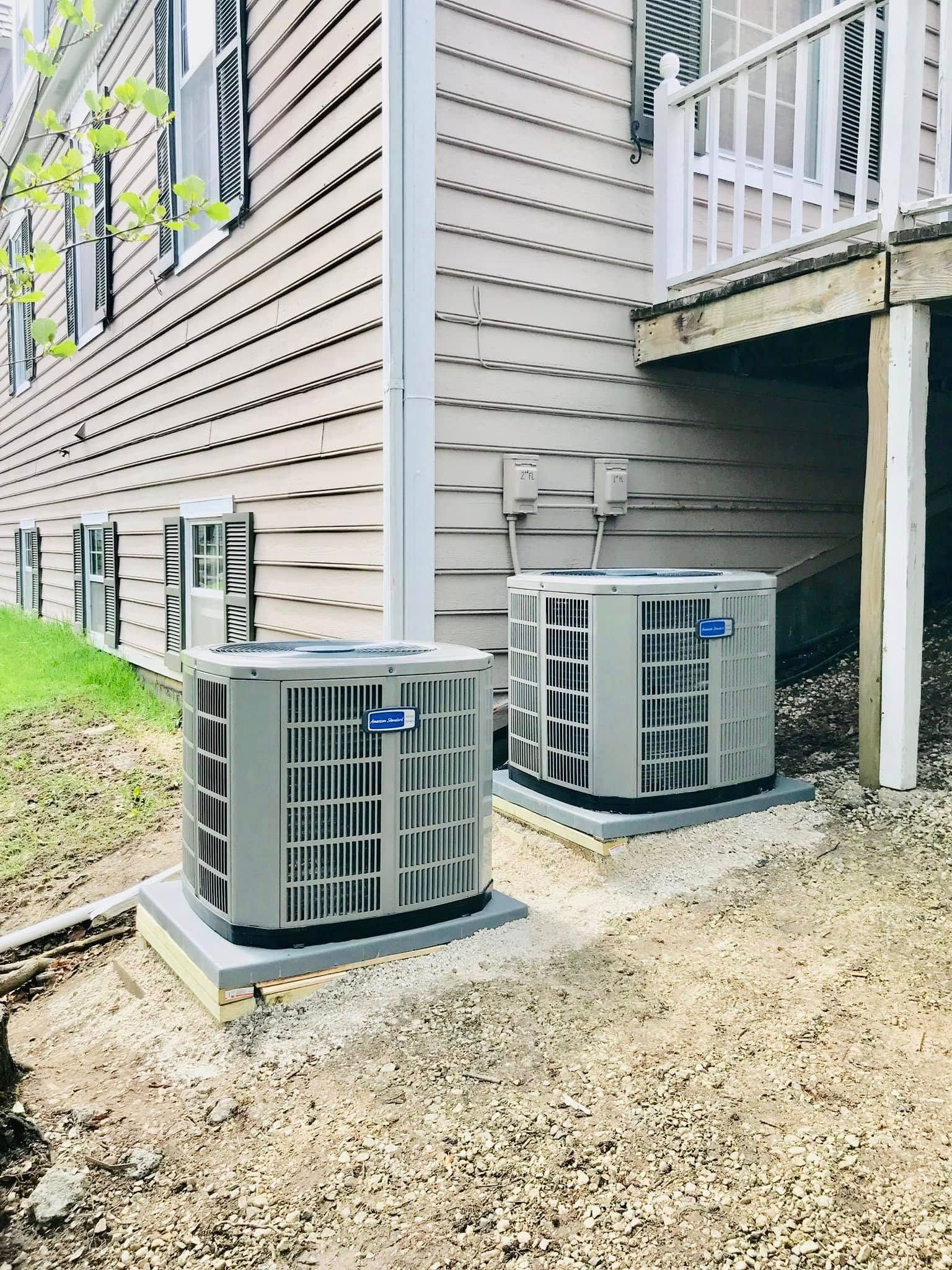 Two outdoor HVAC units beside a gray house under a raised deck