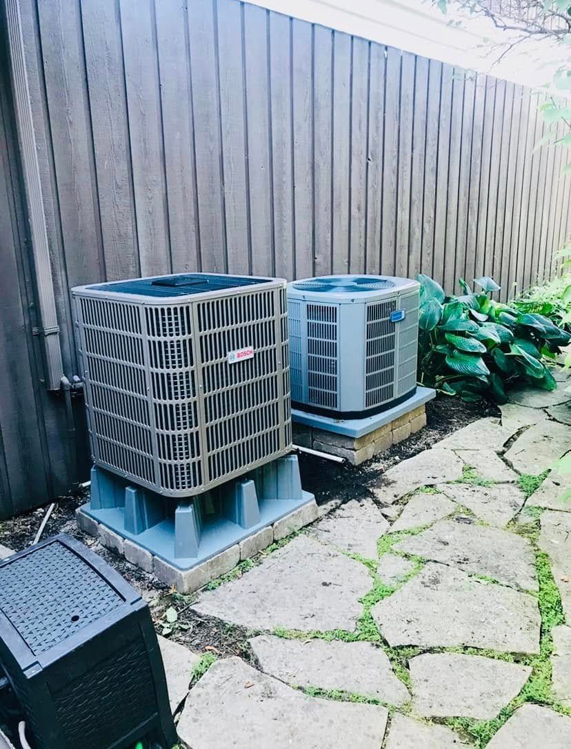 Two outdoor air conditioning units beside a wooden fence on a stone patio, with plants nearby