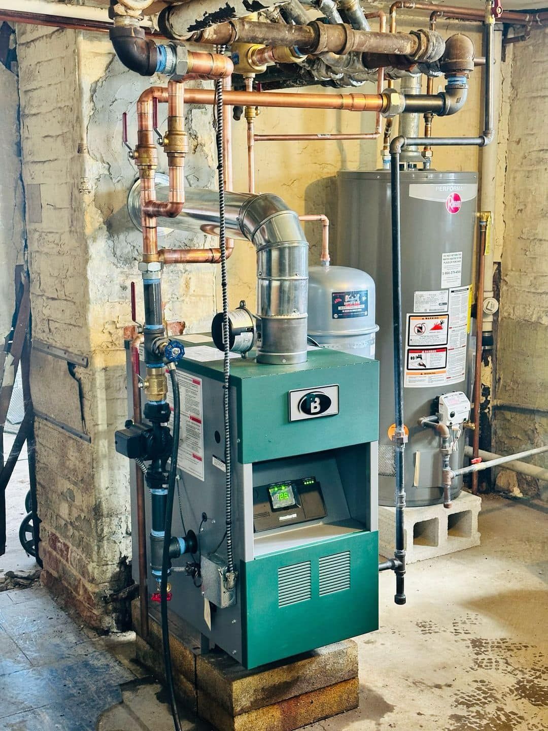 Green boiler furnace and water heater with exposed pipes in a basement utility room.