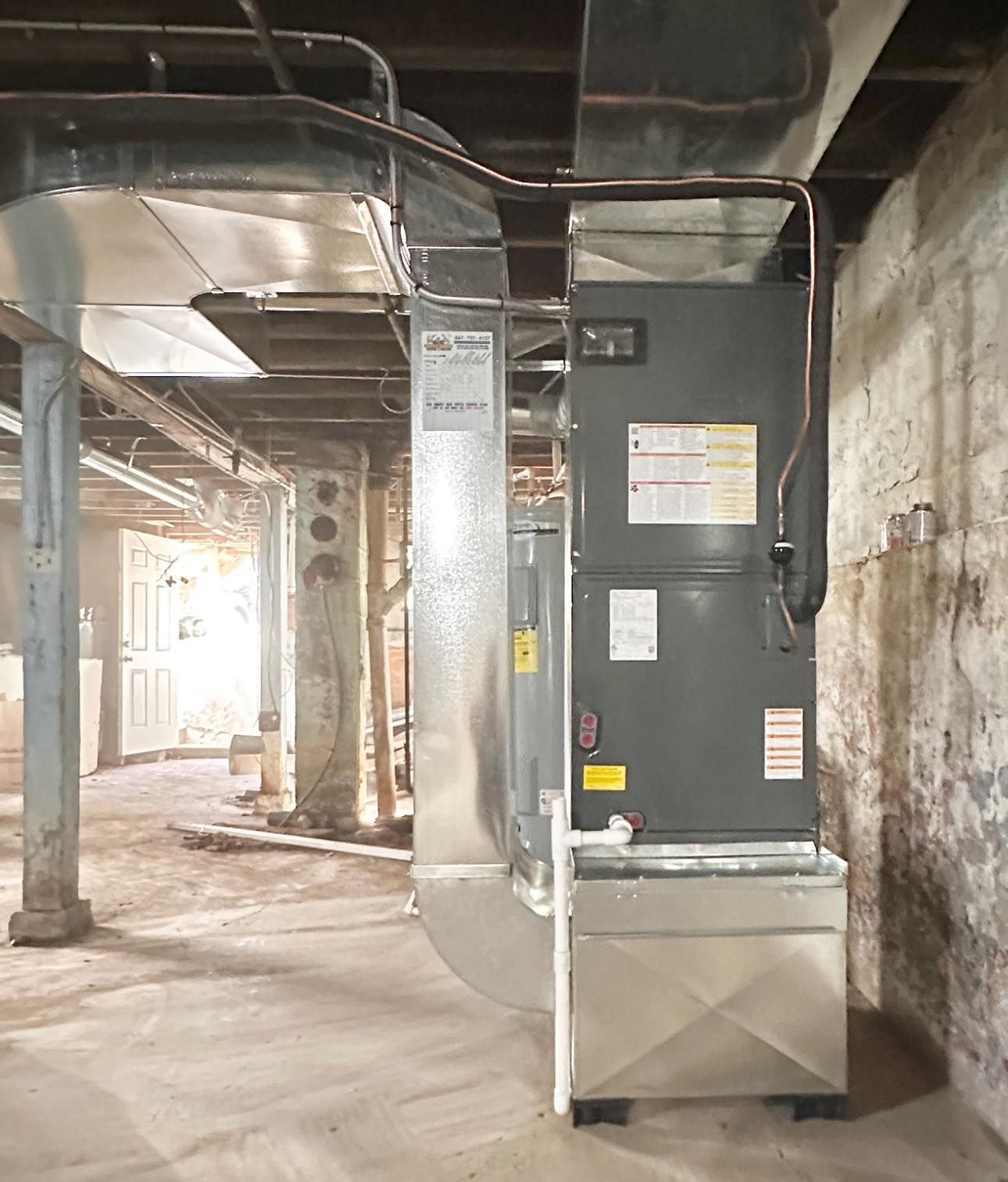 Industrial basement with a large gray HVAC unit, exposed pipes, and concrete walls.