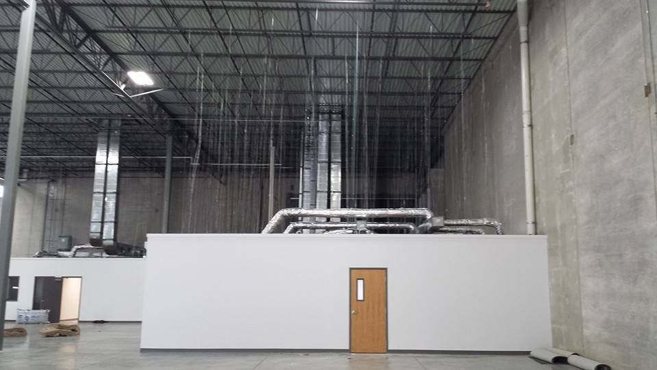 Industrial warehouse interior with a white enclosure and wooden door beneath exposed ceiling pipes.