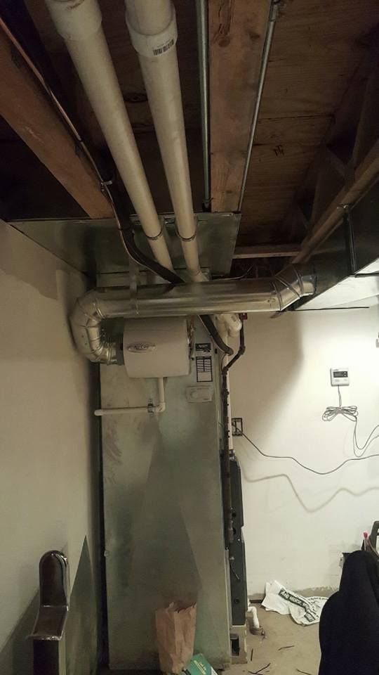 Basement utility room with exposed pipes, ductwork, and hanging wires along a concrete wall.